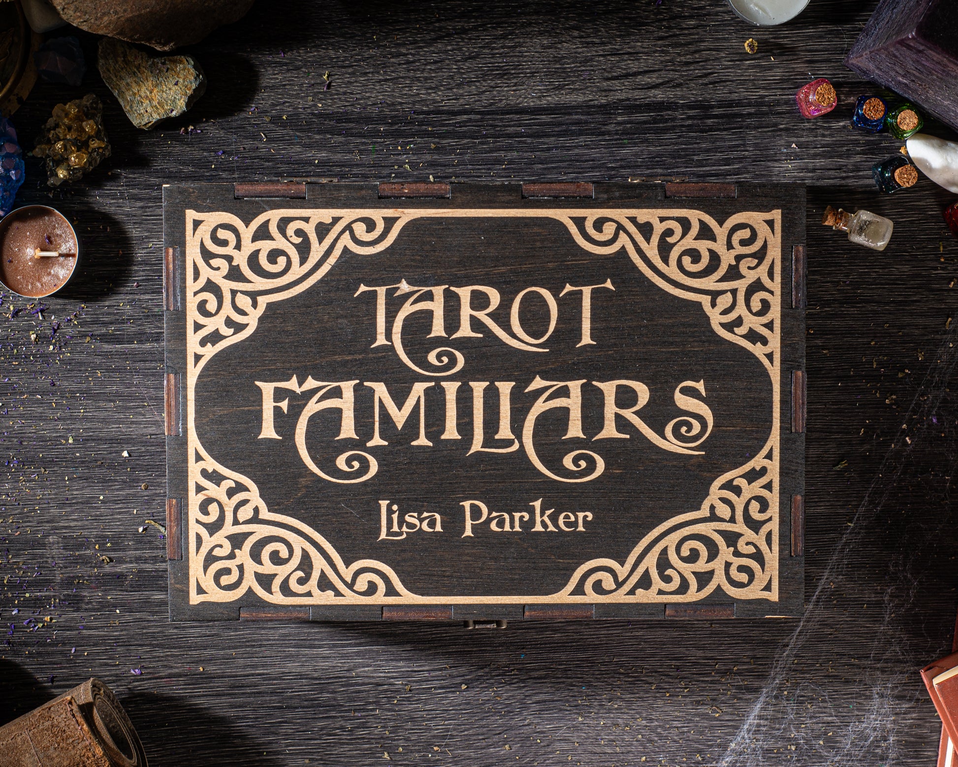 A wooden box with a decorative border and the text "Tarot Familiars" on it, along with the name "Lisa Parker" below. The box is placed on a wooden surface, surrounded by various objects such as rocks, candles, and other items.