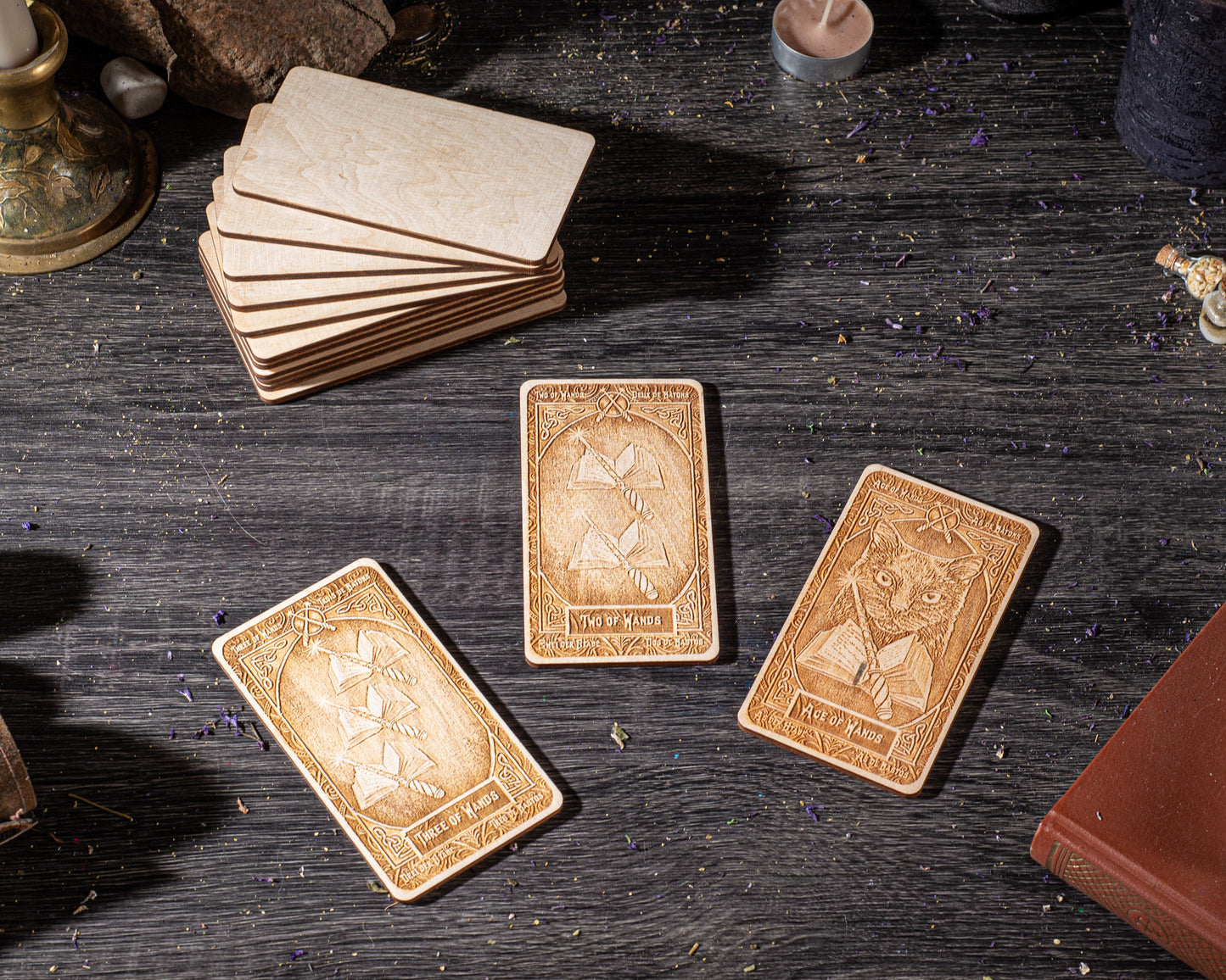 three wooden tarot cards with intricate designs, placed on a dark wooden surface alongside other objects such as a candle and a book.
