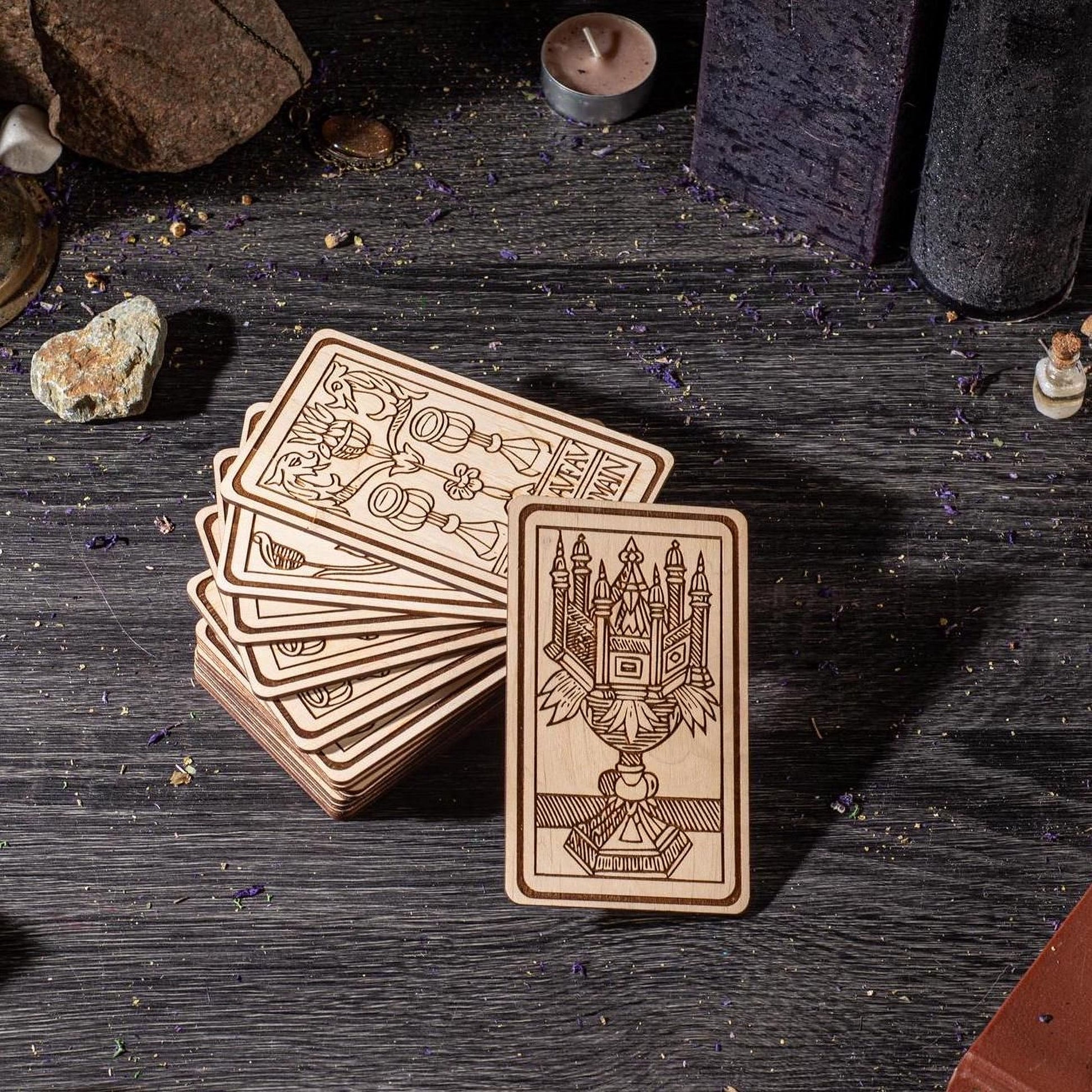A collection of wooden tarot cards is displayed on a dark wooden surface, with various objects such as candles, rocks, and crystals scattered around.