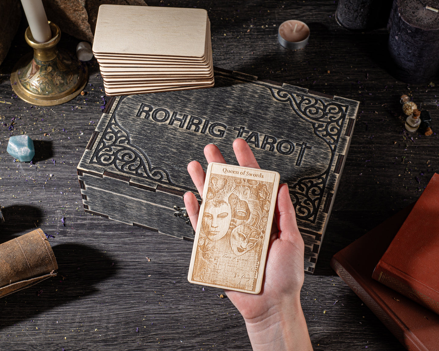 A hand holding a wooden tarot card with intricate designs, surrounded by various objects such as a stack of cards, a wooden box, and other items on a dark wooden surface.