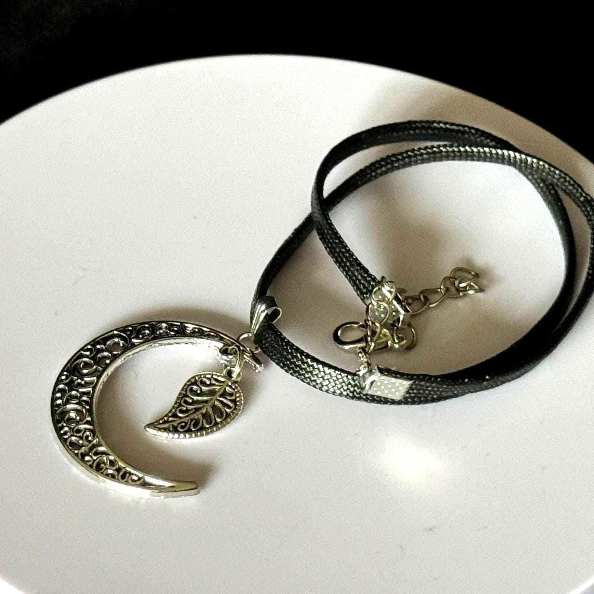 A silver necklace with a pendant featuring a crescent moon and leaf design, resting on a white surface.