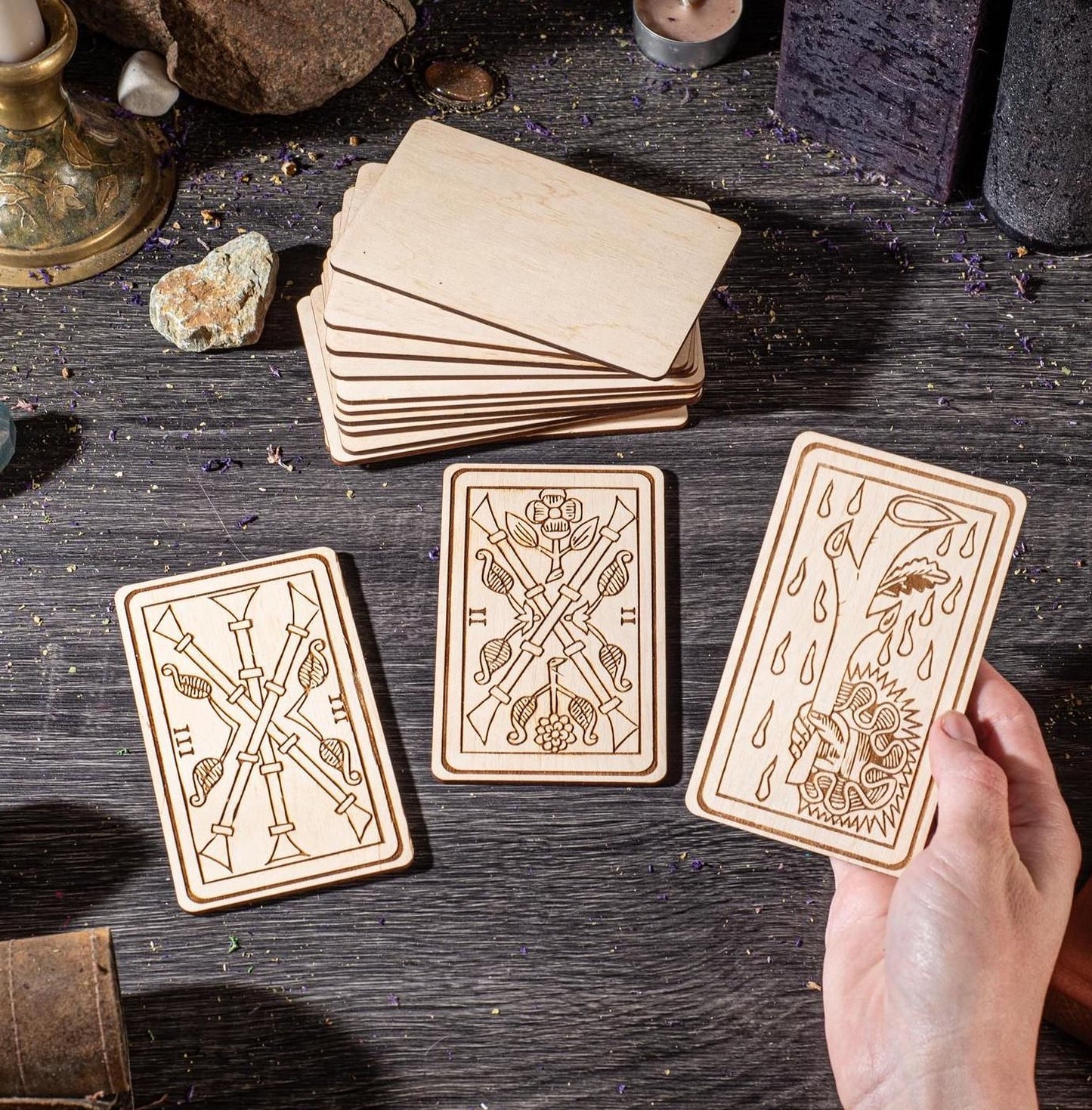 a person's hands holding three wooden tarot cards, each with intricate designs, on a dark wooden surface. The cards are arranged in a row, and there are additional wooden cards and objects in the background.