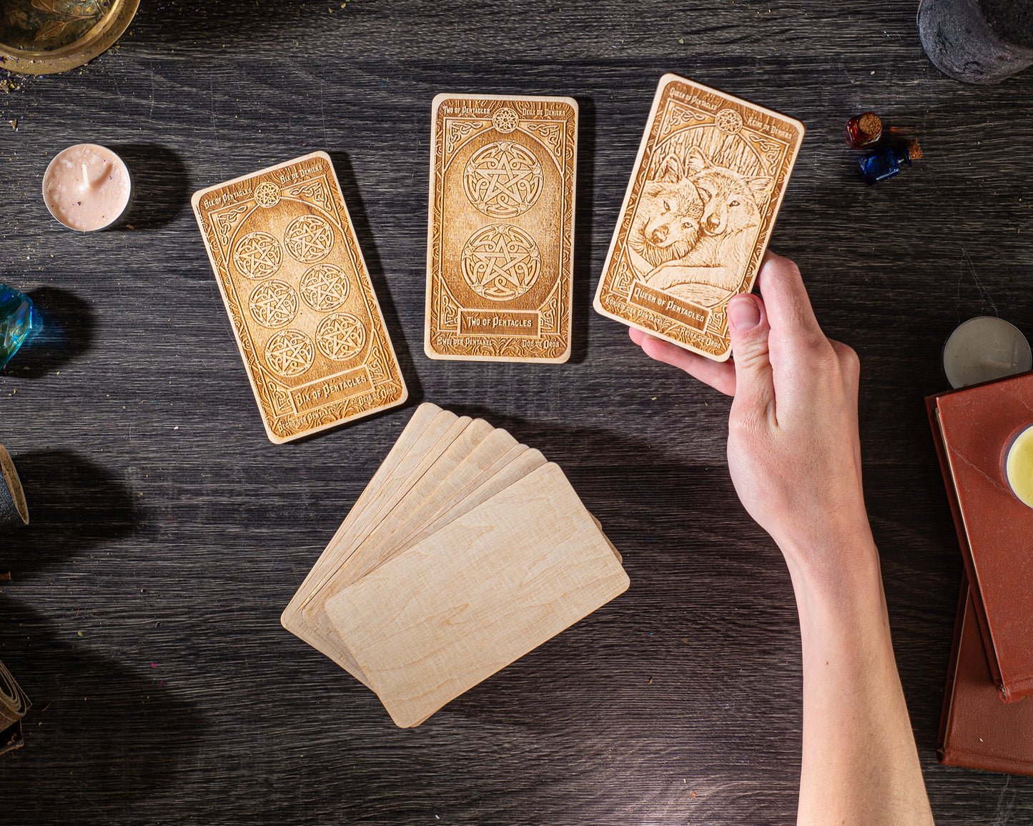 a hand holding a wooden card with a wolf design, surrounded by other wooden cards and various objects on a dark wooden surface.