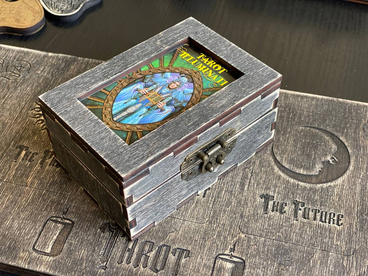 A wooden box with a metal clasp, featuring an illustration of a woman with a blue and green background. The box is placed on a wooden surface with embossed designs.