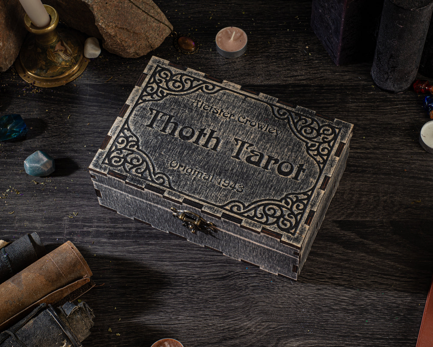 A wooden box with a decorative border sits on a wooden surface, surrounded by various objects such as candles, gemstones, and books.