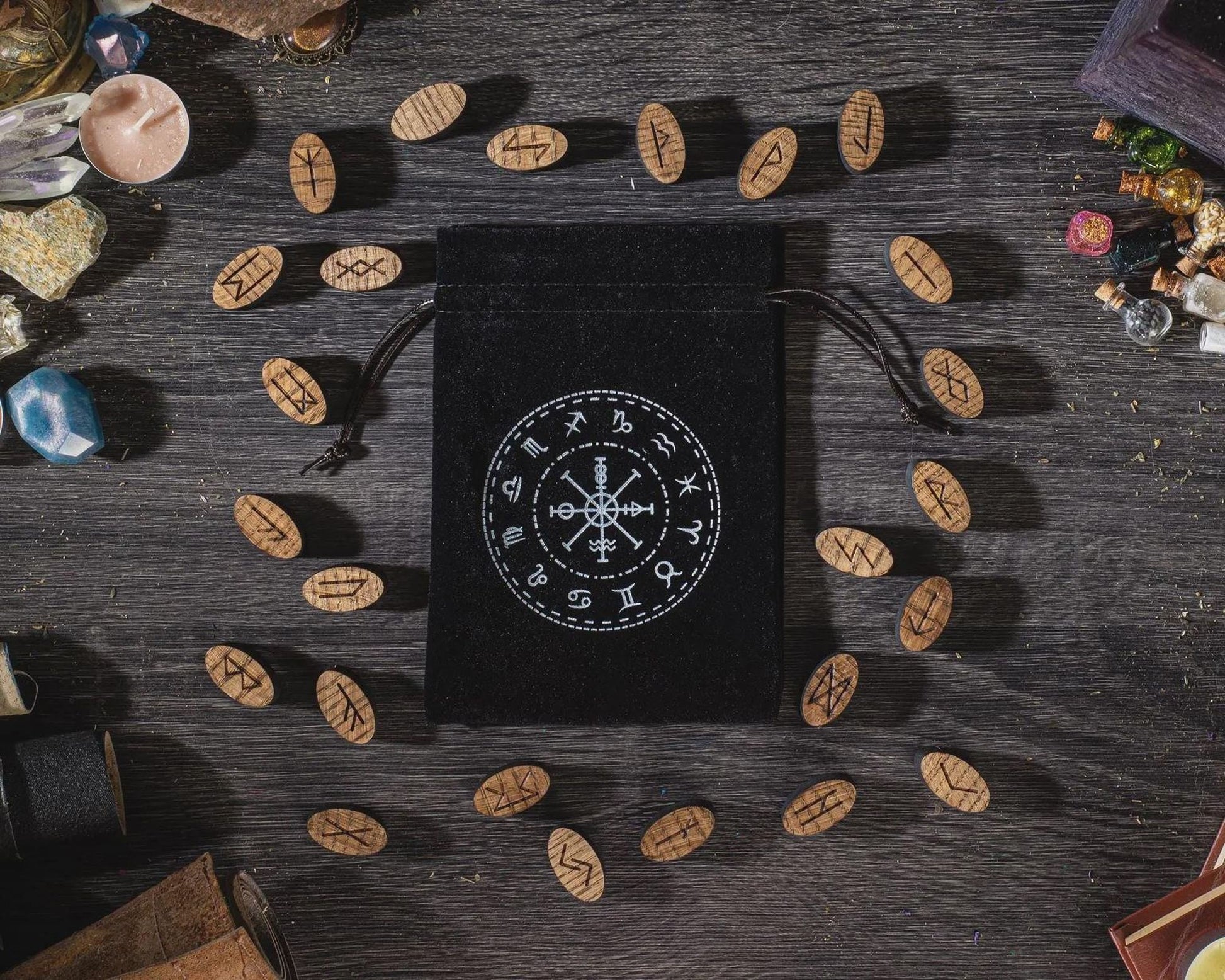 a black pouch with a white design on it, surrounded by various objects such as crystals, stones, and wooden pieces on a wooden surface.