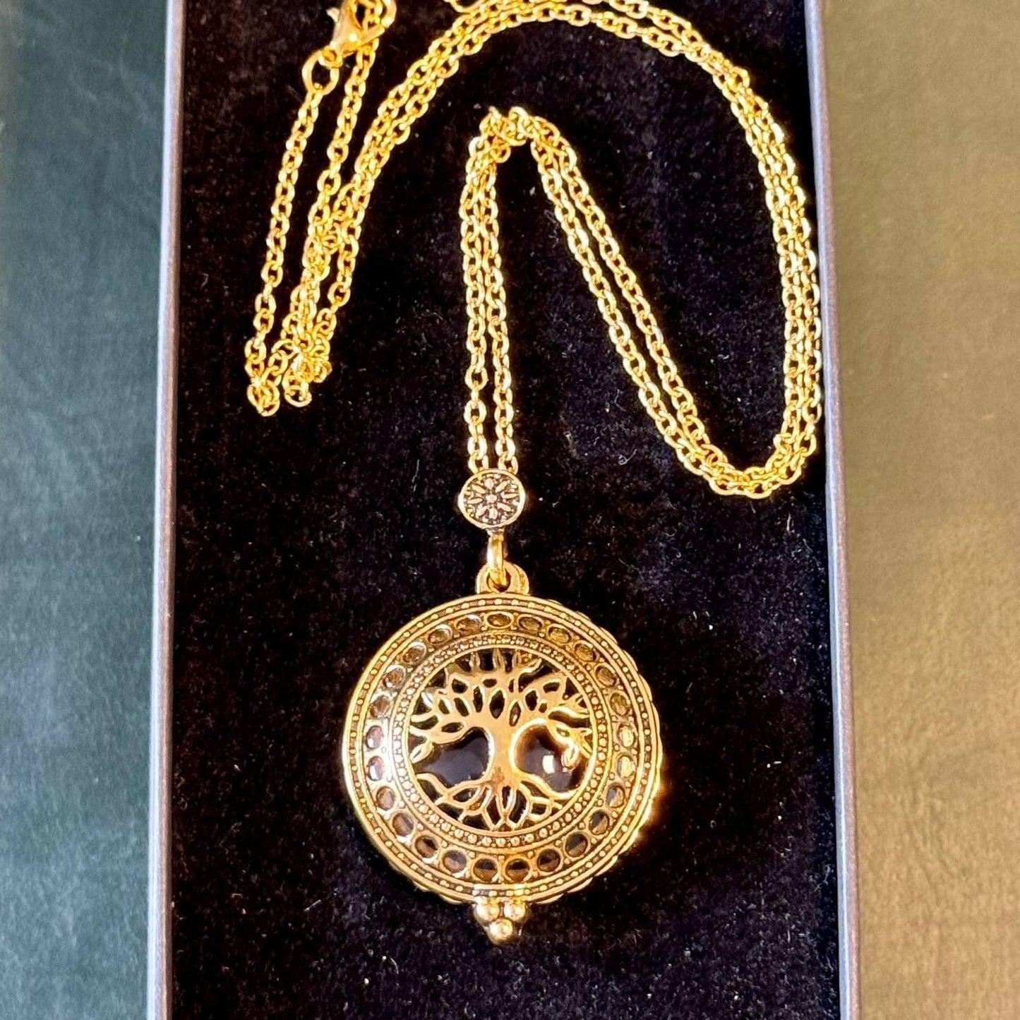 A gold-colored necklace with a circular pendant featuring a tree design, displayed in a black velvet box.