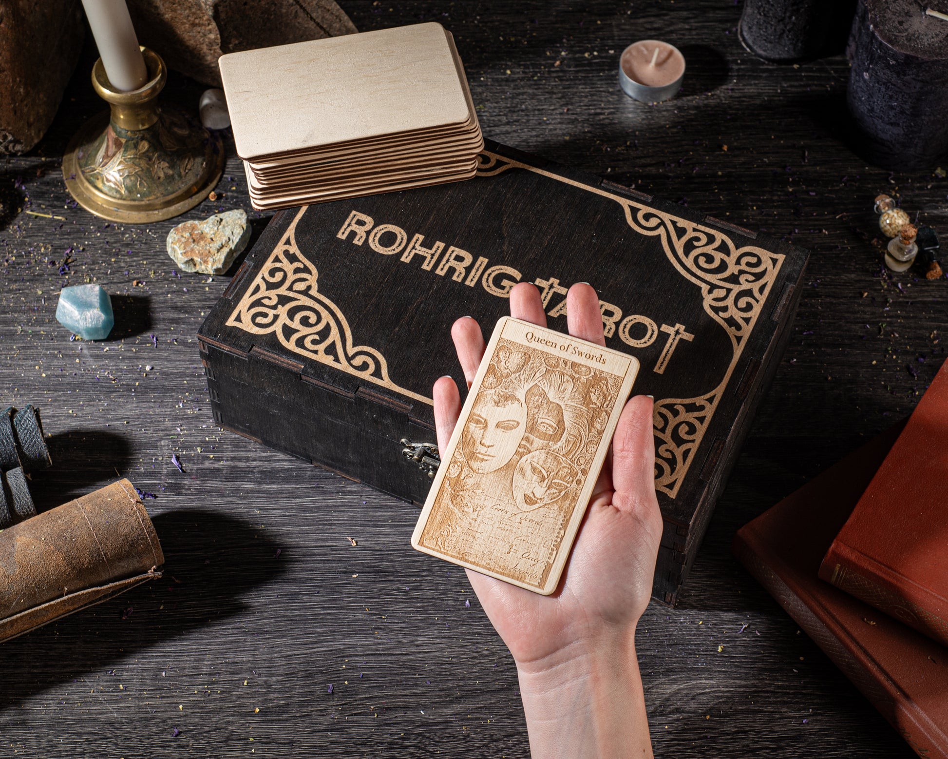 A hand holding a wooden tarot card with the name "ROHRIGOT" on it, surrounded by various objects such as a stack of cards, a candle, and a book.