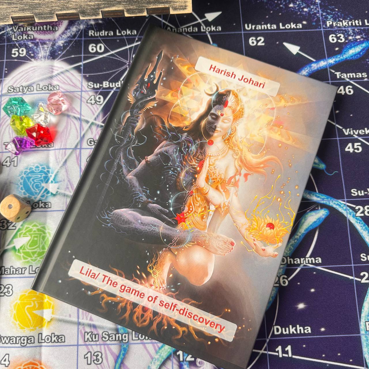 a book titled "Lila/The game of self-discovery" with a colorful, artistic cover depicting a woman with a golden head and a black body. The book is placed on a board game with various game pieces and symbols scattered around it.