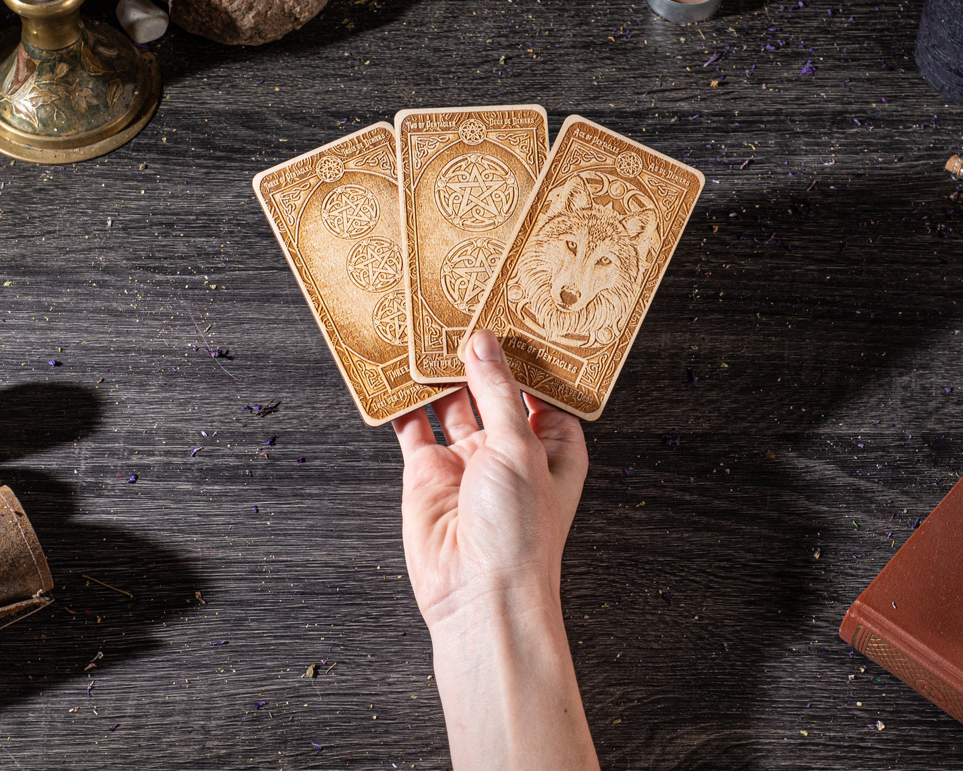 A hand holding three wooden tarot cards with intricate designs on them, placed on a dark wooden surface.