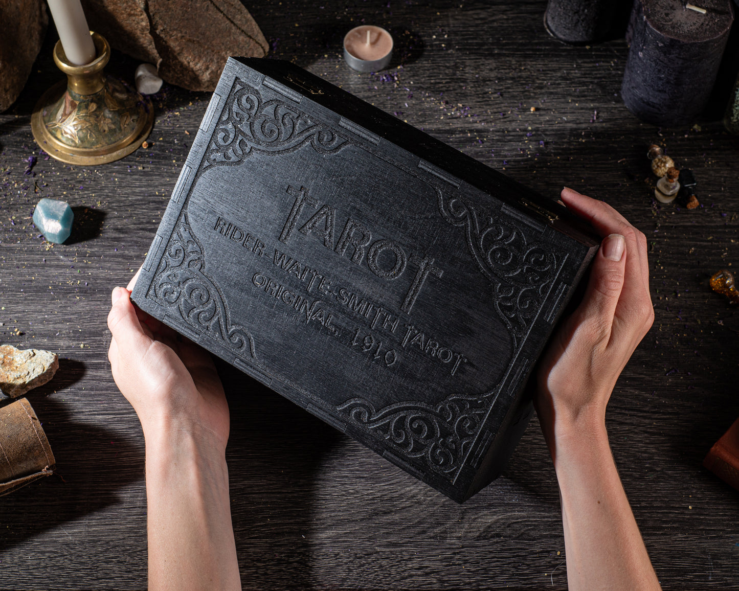 A person's hands are holding a black tarot card box with ornate designs on it, placed on a wooden surface with various objects around it.