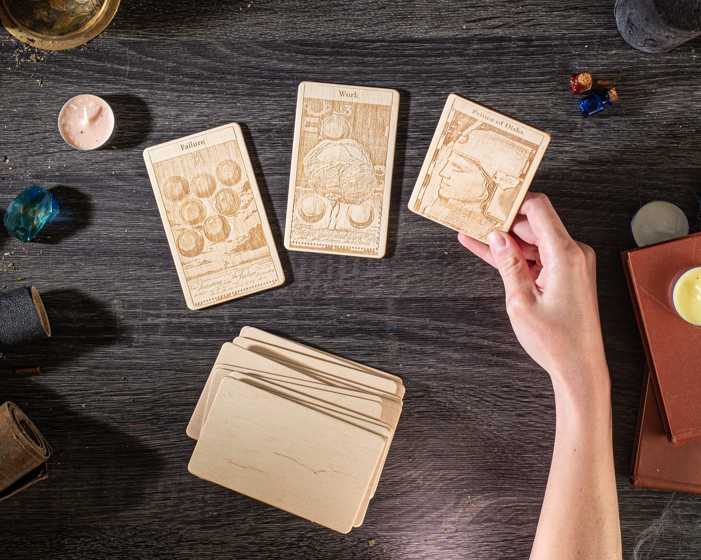 a hand holding a wooden card with a picture of a person on it, surrounded by various objects such as candles, books, and other wooden items on a dark wooden surface.