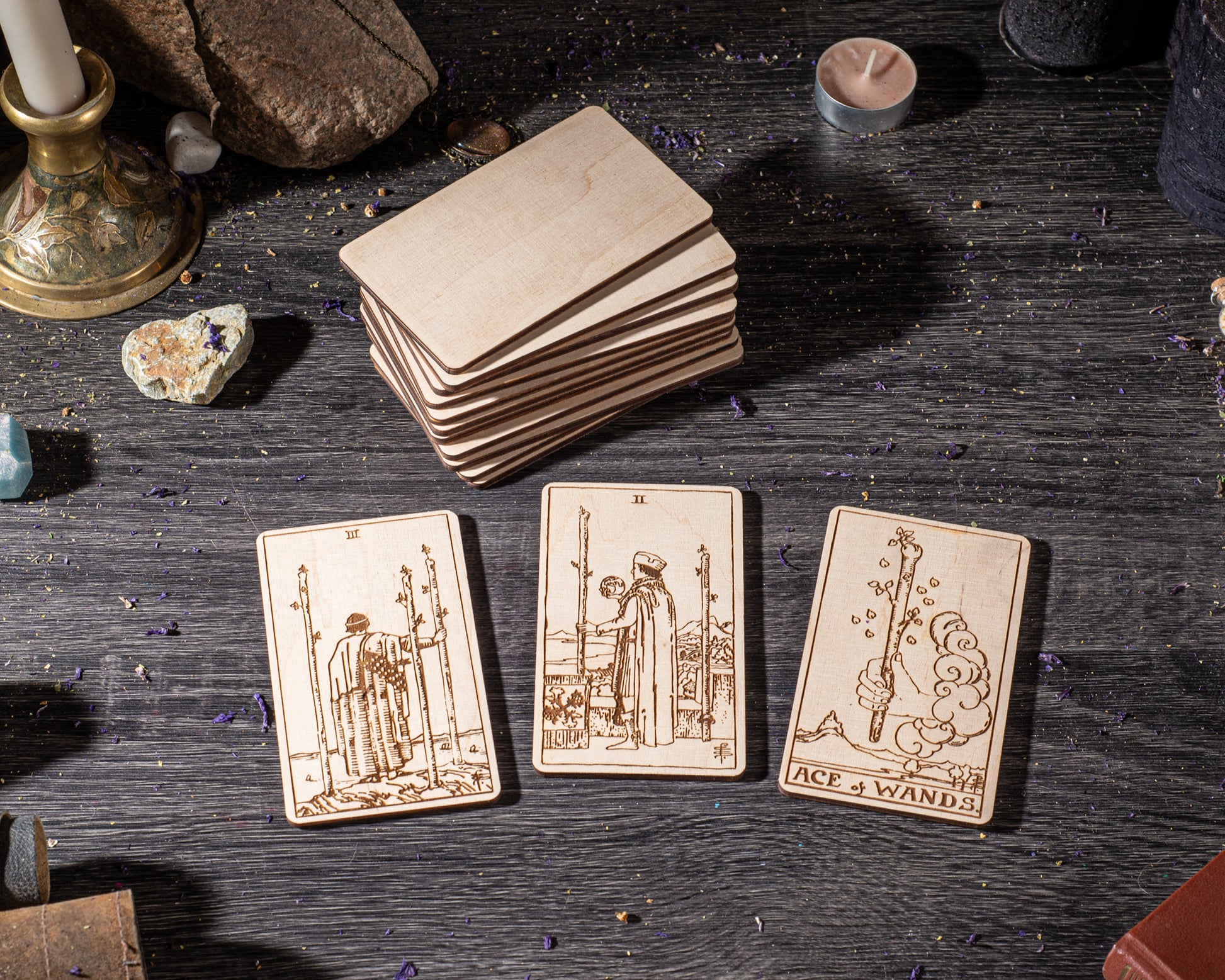 three wooden tarot cards with illustrations, placed on a dark wooden surface. There are also various objects such as a candle, a rock, and some crystals in the background.
