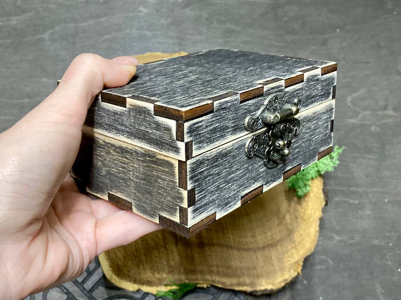 A hand is holding a small, wooden box with a metal clasp on top.