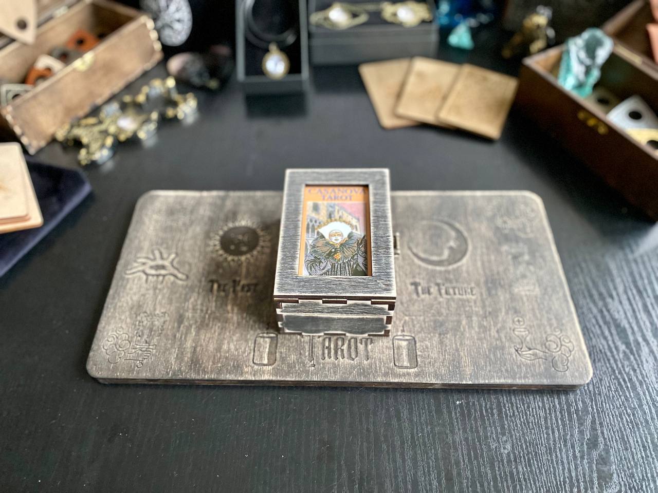 a wooden box with a metal lid, placed on a wooden surface. The box has a label that reads "T-T-T-T-T" and "T-T-T-T" is visible on the lid. The background includes various objects such as a clock, a necklace, and a small figurine.