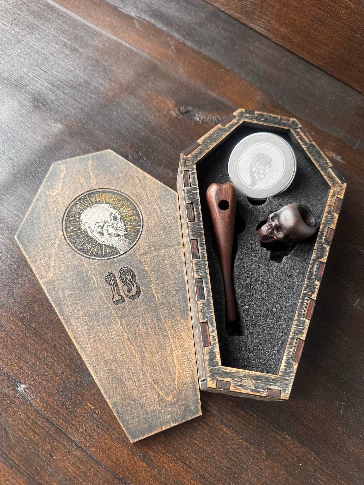A wooden box with a skull design on it, containing a wooden pipe and a small metal object, placed on a wooden surface.