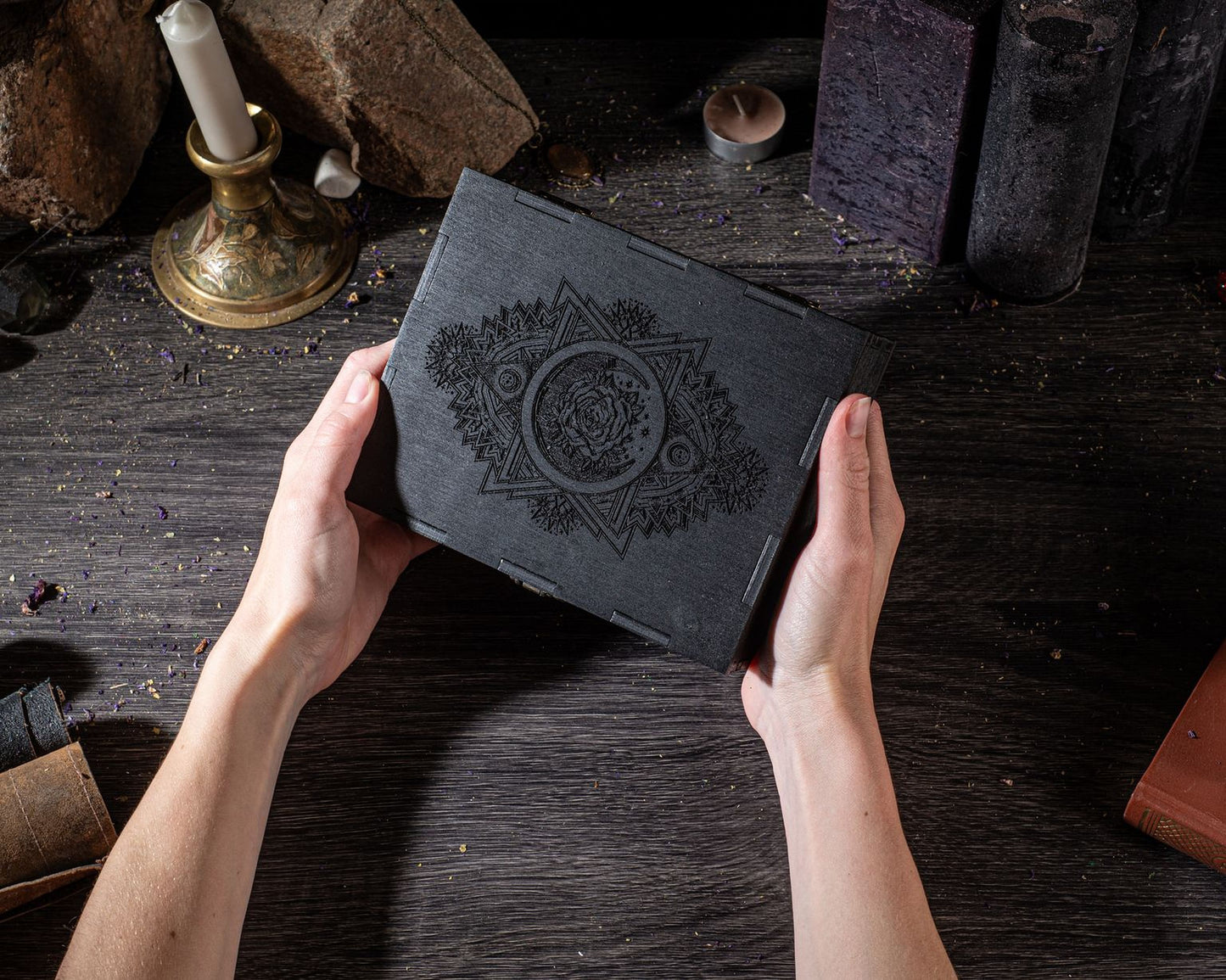 A pair of hands holding a black book with an intricate design on the cover, placed on a wooden surface with various objects in the background.