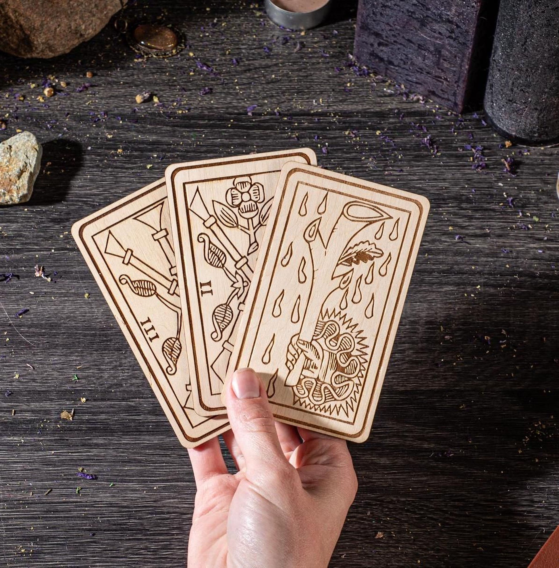 A hand is holding three wooden cards with intricate designs on them, placed on a wooden surface with scattered objects around.