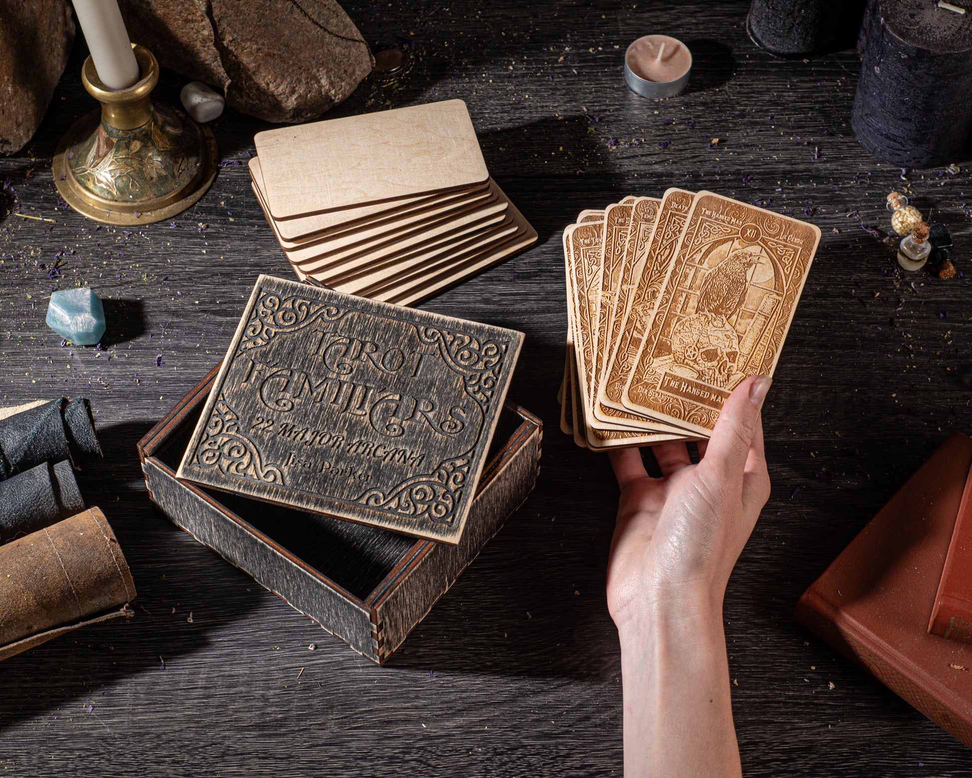 a hand holding a deck of playing cards, with a wooden box and other items such as candles and books visible on a dark wooden surface.