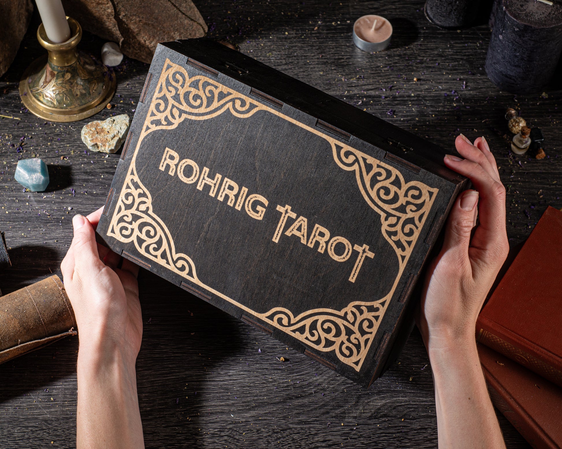A person's hands holding a black box with gold decorative borders, which has the word "Rohrig Tarot" written on it. The box is placed on a wooden surface, surrounded by various objects such as books, crystals, and a candle.