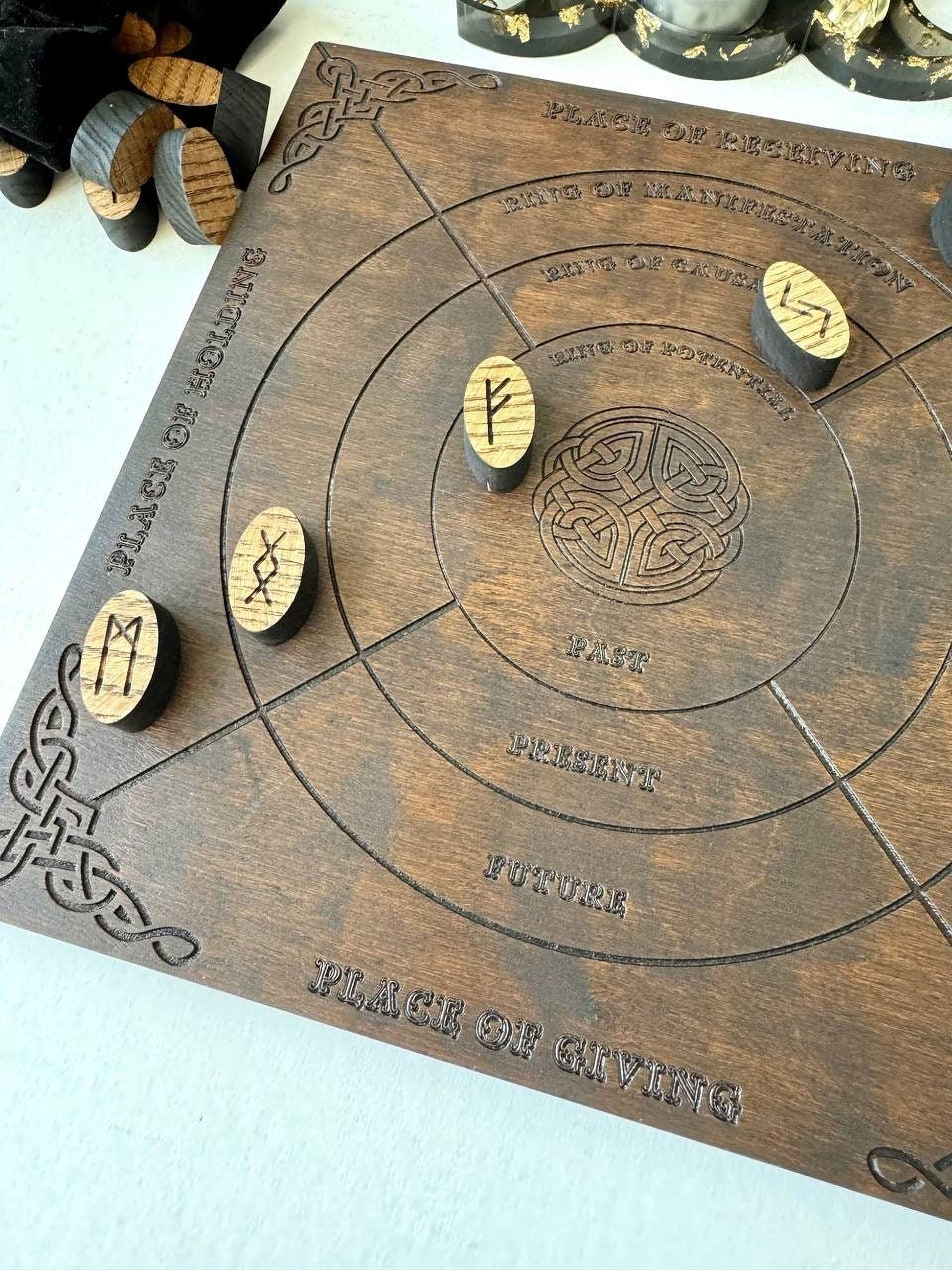 a wooden board with various carved symbols and text, including the words "Place of Reserving", "Place of Magic", "Place of Giving", and "Place of Death". There are also several wooden objects, such as a cork and a piece of driftwood, scattered around the board.