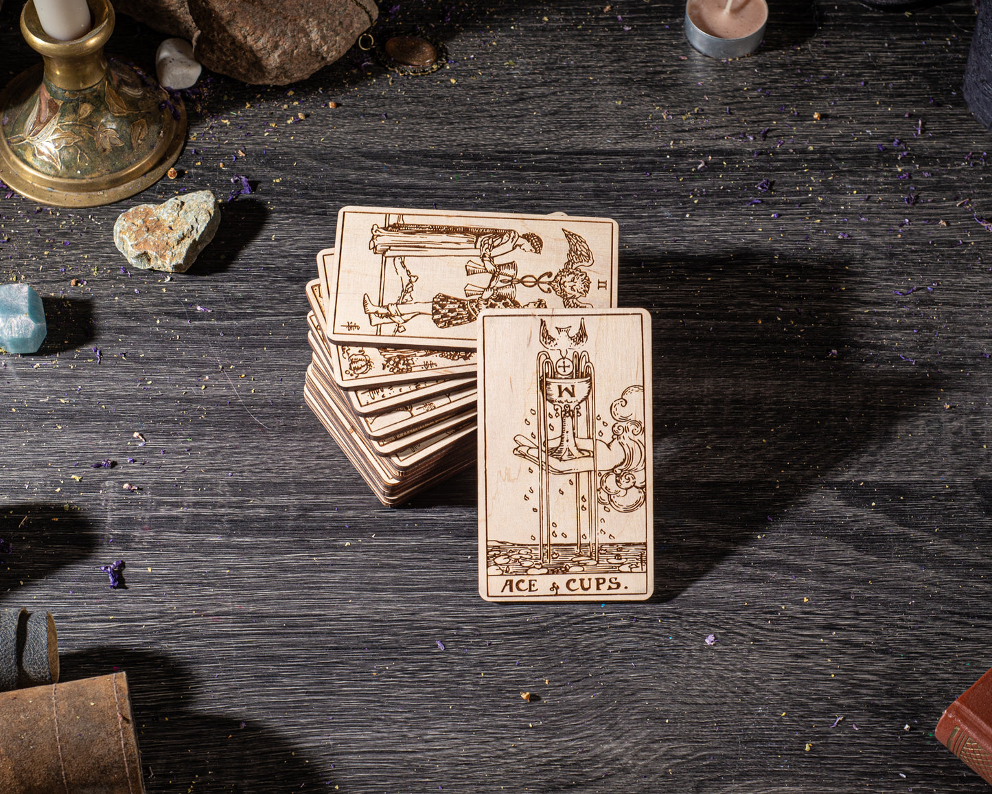 A stack of wooden tarot cards is displayed on a wooden surface, with various objects such as crystals, a candle, and a book in the background.