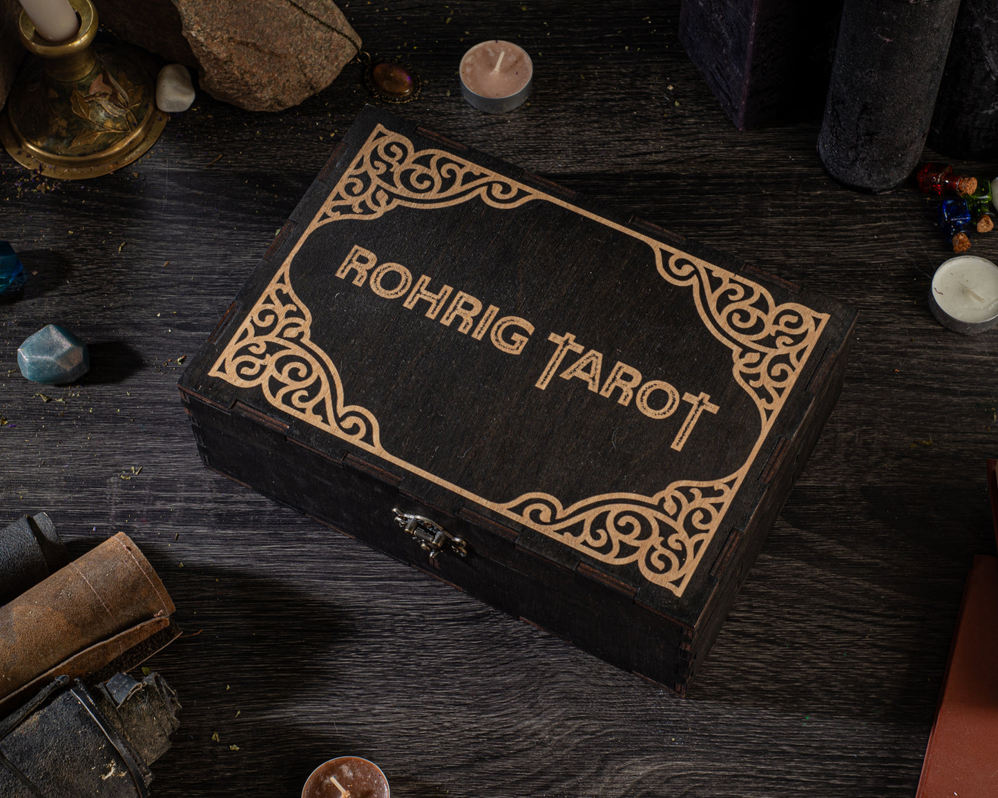A wooden box with the name "Rohrig Tarot" embossed on it, surrounded by various objects such as candles, gemstones, and books on a dark wooden surface.