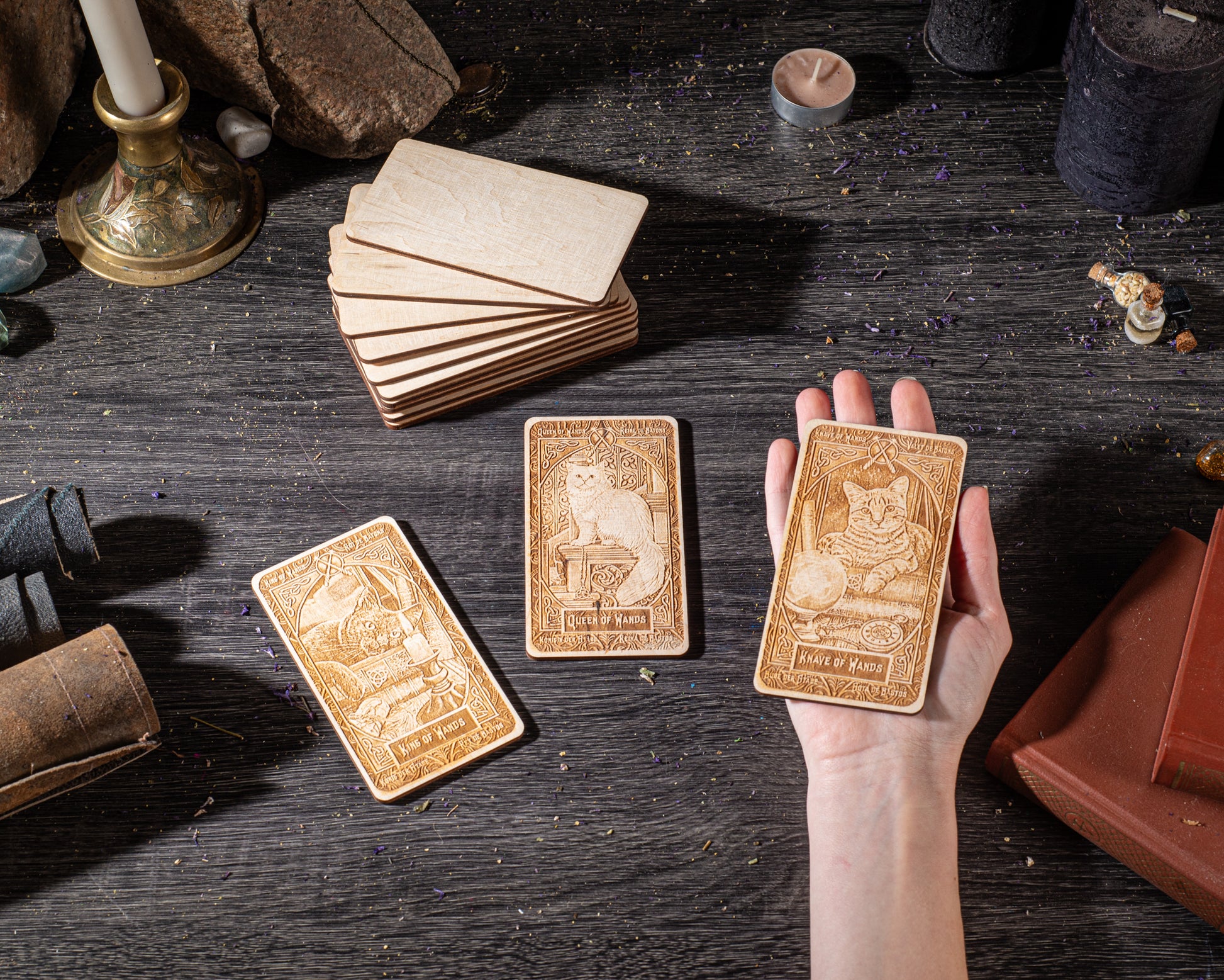 A hand is holding a wooden card with an illustration of a cat, surrounded by other wooden cards and objects on a dark wooden surface.