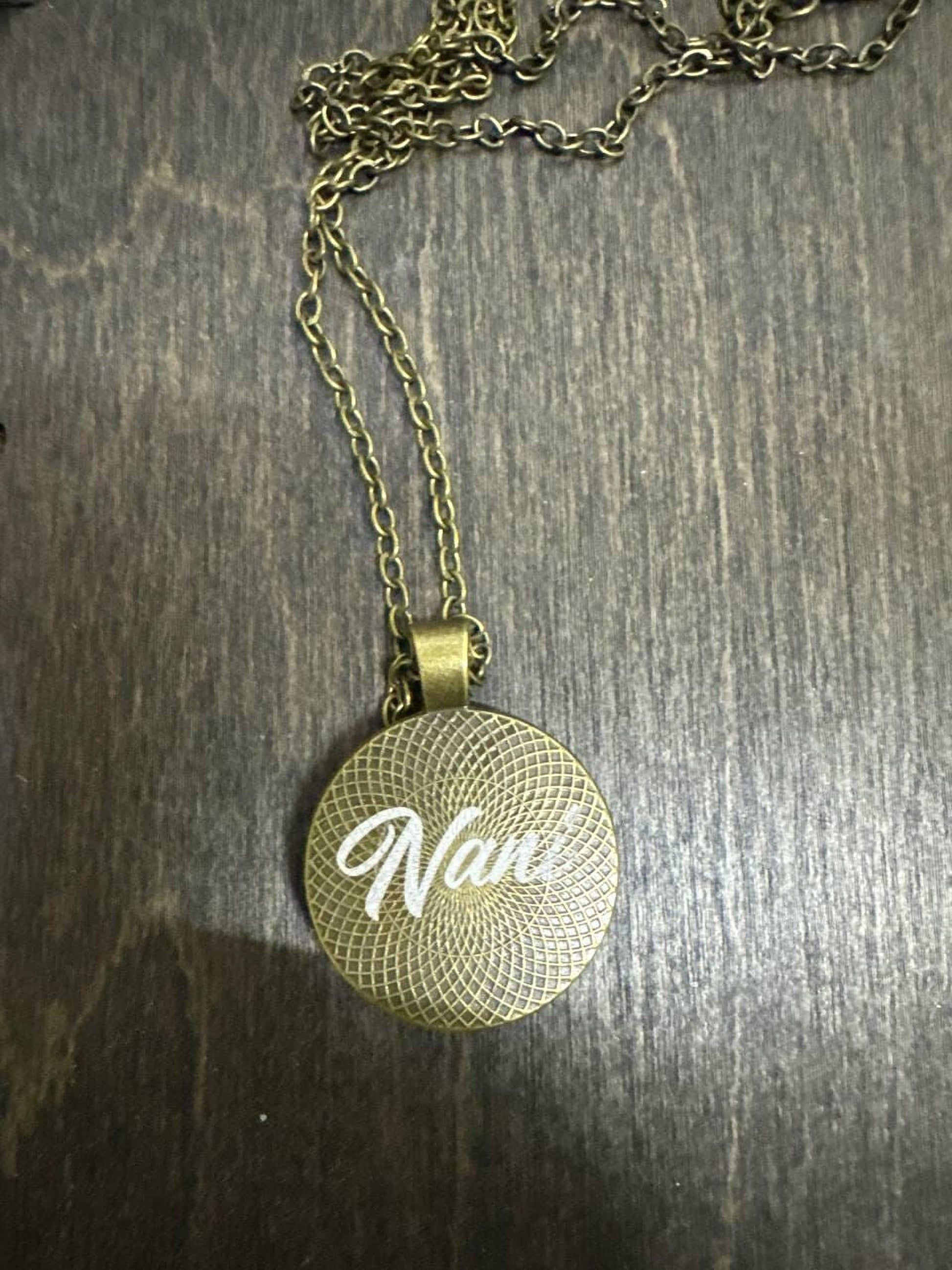 A gold-colored necklace with a circular pendant featuring the word "Nani" engraved on it, resting on a wooden surface.