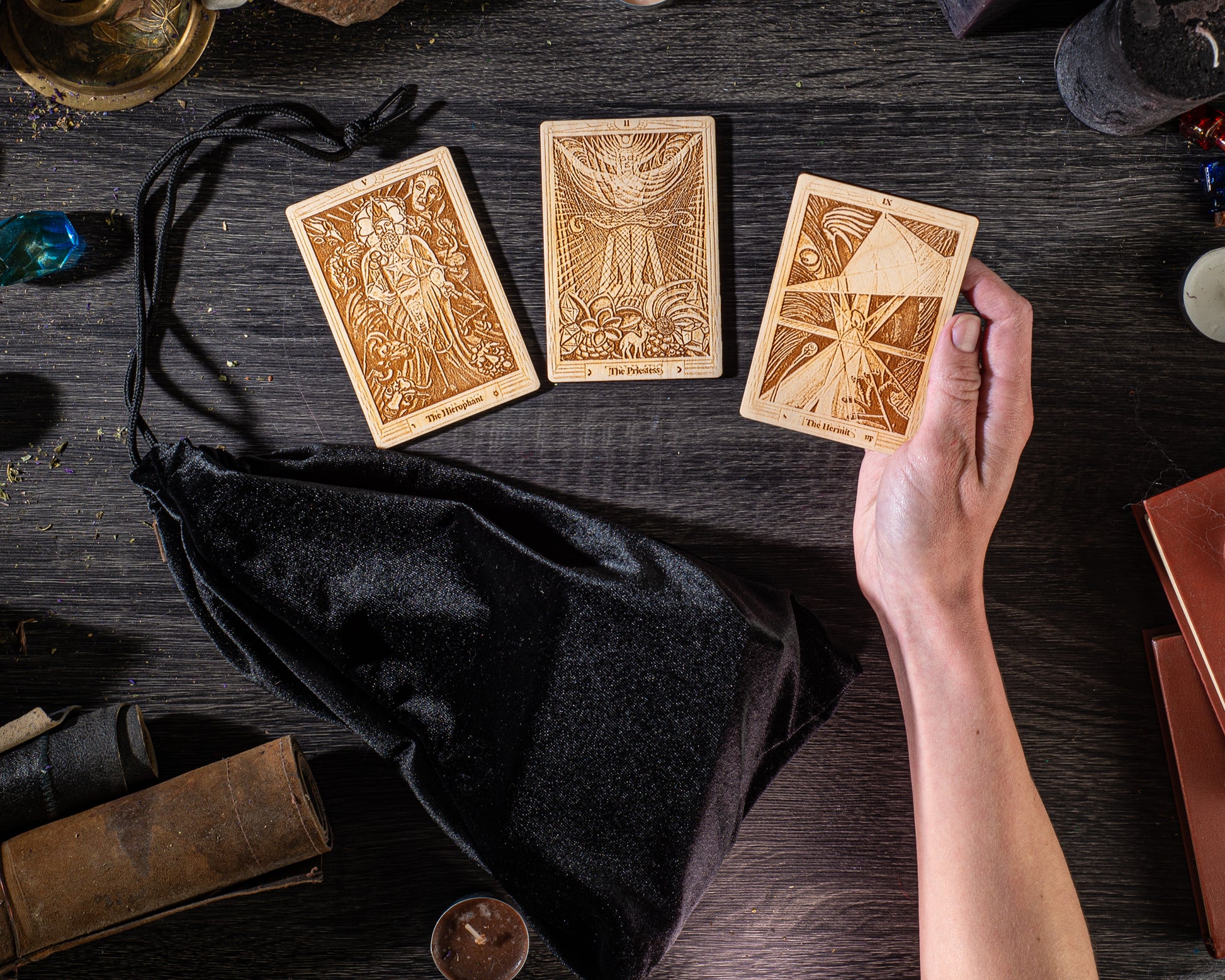 a person's hand holding three wooden tarot cards, with a black bag and various other items on a wooden surface.