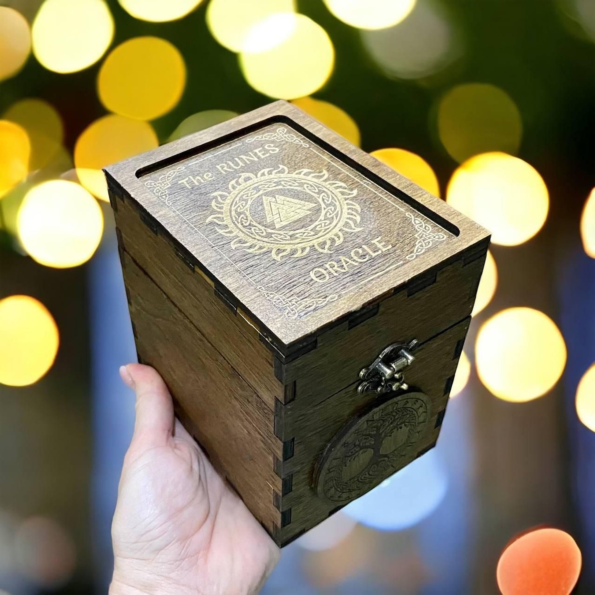 A hand is holding a wooden box with the text "The Runes Oracle" on it, against a blurred background with colorful bokeh lights.