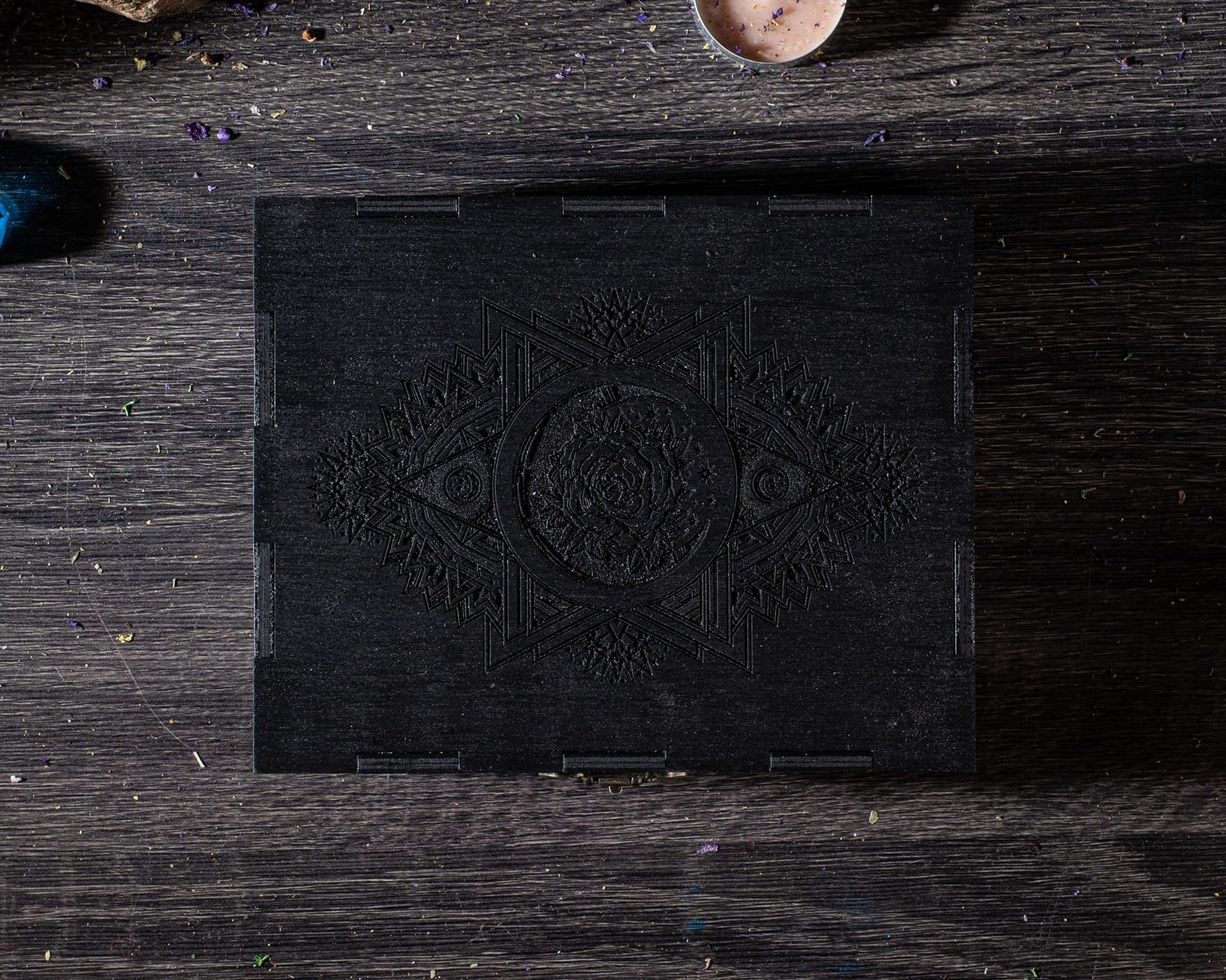 A black wooden box with an intricate design on its surface, placed on a wooden surface with scattered objects around it.