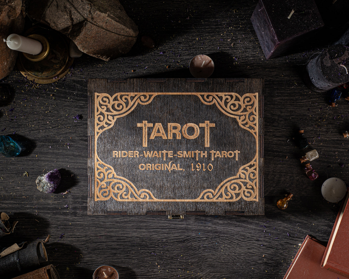 a wooden box with the name "TAROT" and the text "RIDER-WAITE SMITH TAROT ORIGINAL. 1910" on it, surrounded by various objects such as candles, crystals, and books on a dark wooden surface.
