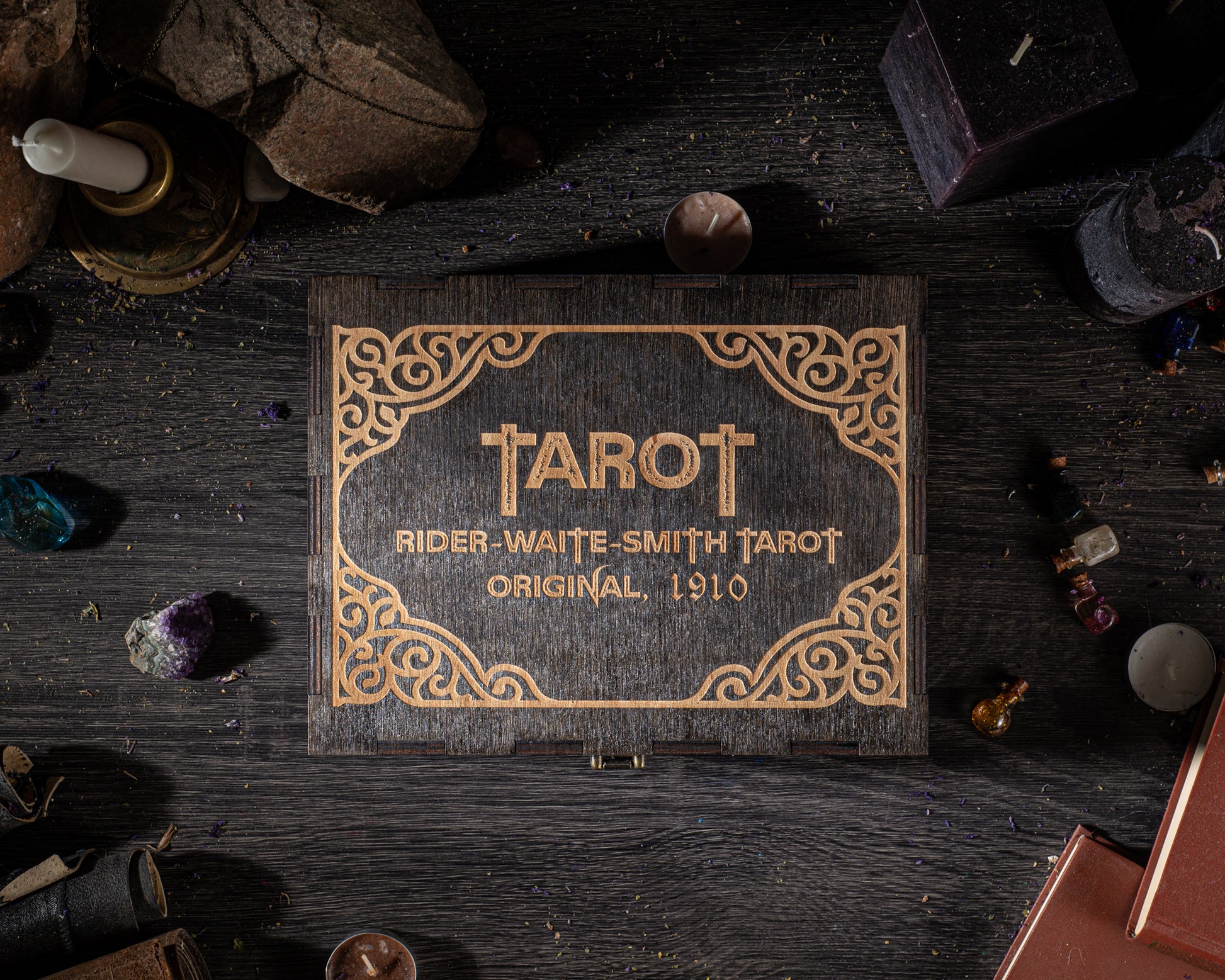 a wooden box with the name "TAROT" and the text "RIDER-WAITE SMITH TAROT ORIGINAL. 1910" on it, surrounded by various objects such as candles, crystals, and books on a dark wooden surface.