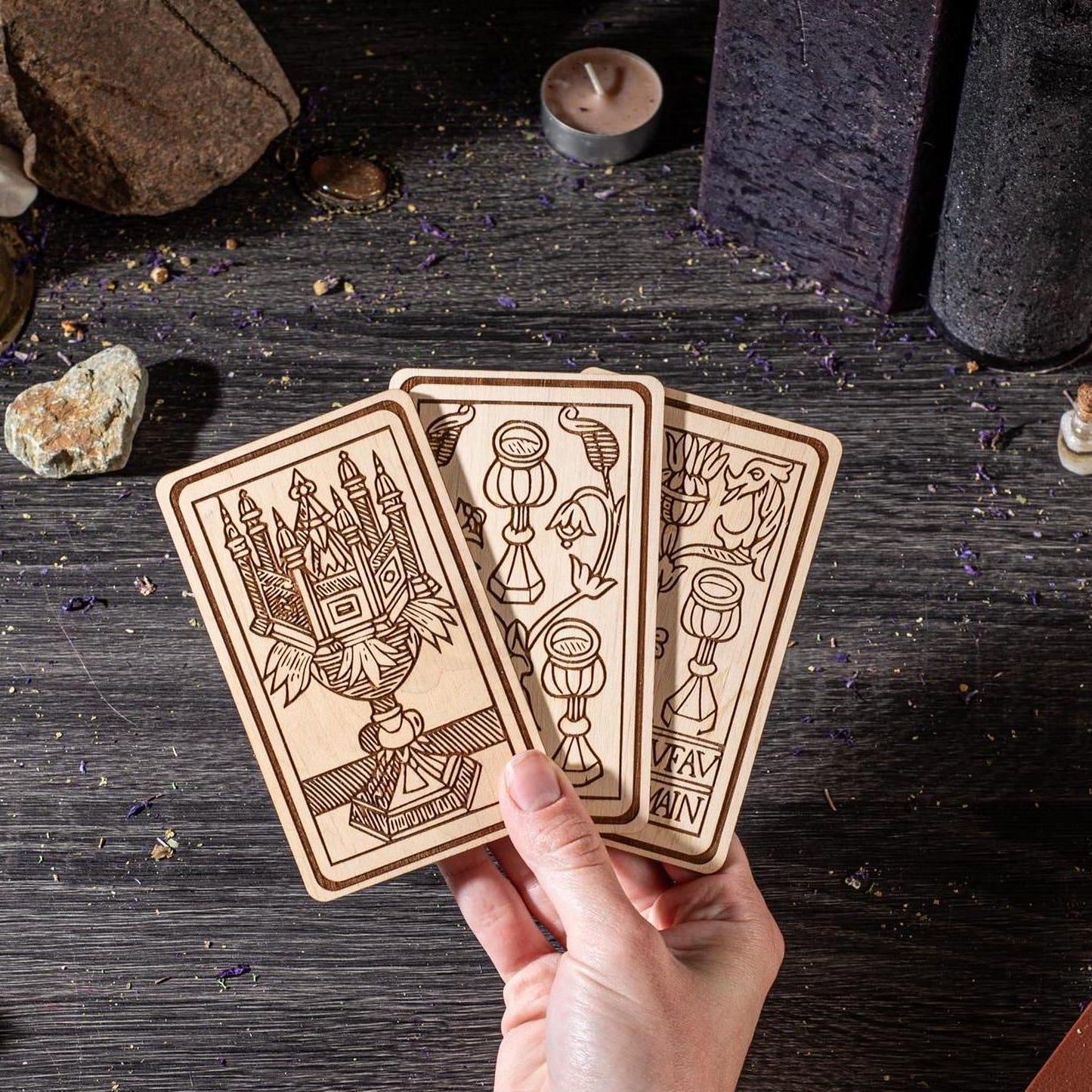 A hand is holding three wooden tarot cards against a dark background, with various objects such as rocks and candles in the background.