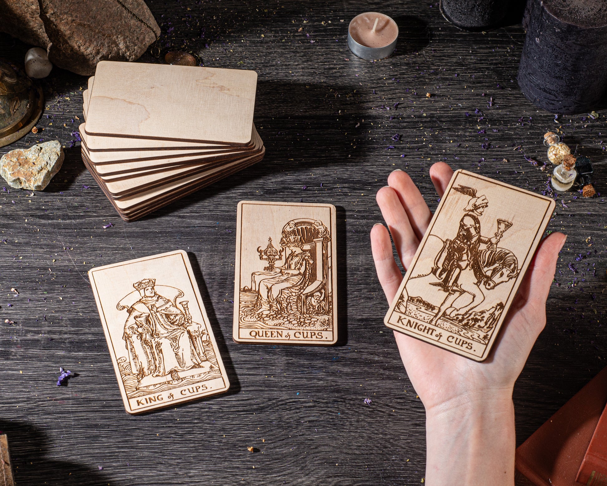 A hand is holding three wooden tarot cards on a dark wooden surface, surrounded by various objects such as candles, crystals, and rocks.
