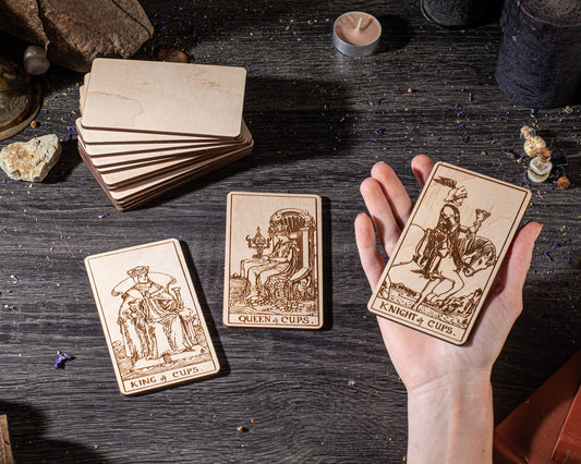 A hand is holding three wooden tarot cards on a dark wooden surface, surrounded by various objects such as candles, crystals, and rocks.