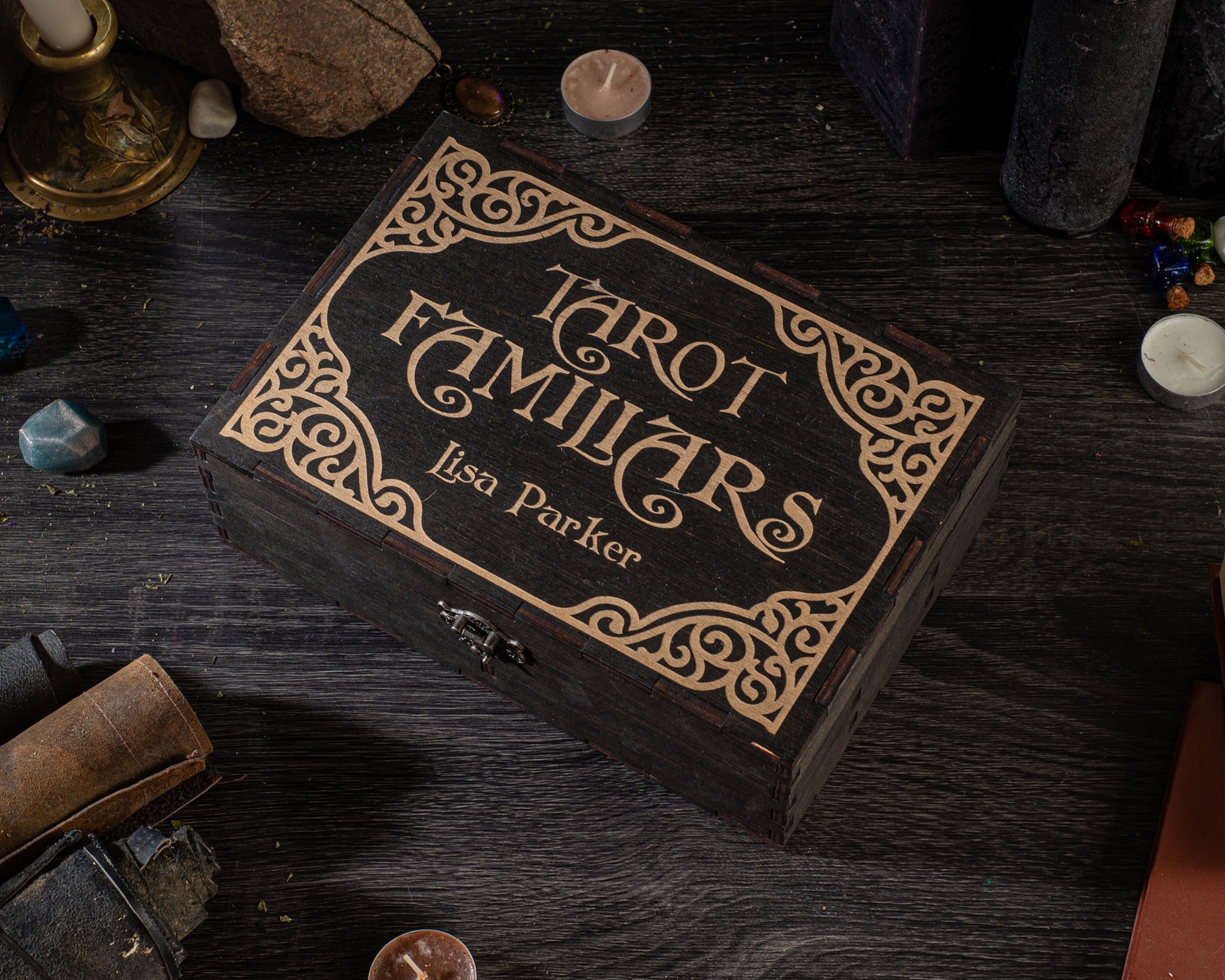 A wooden box with a decorative border, labeled "TURT FAMILIARS Lisa Parker", sits on a dark wooden surface surrounded by various objects such as candles, books, and crystals.