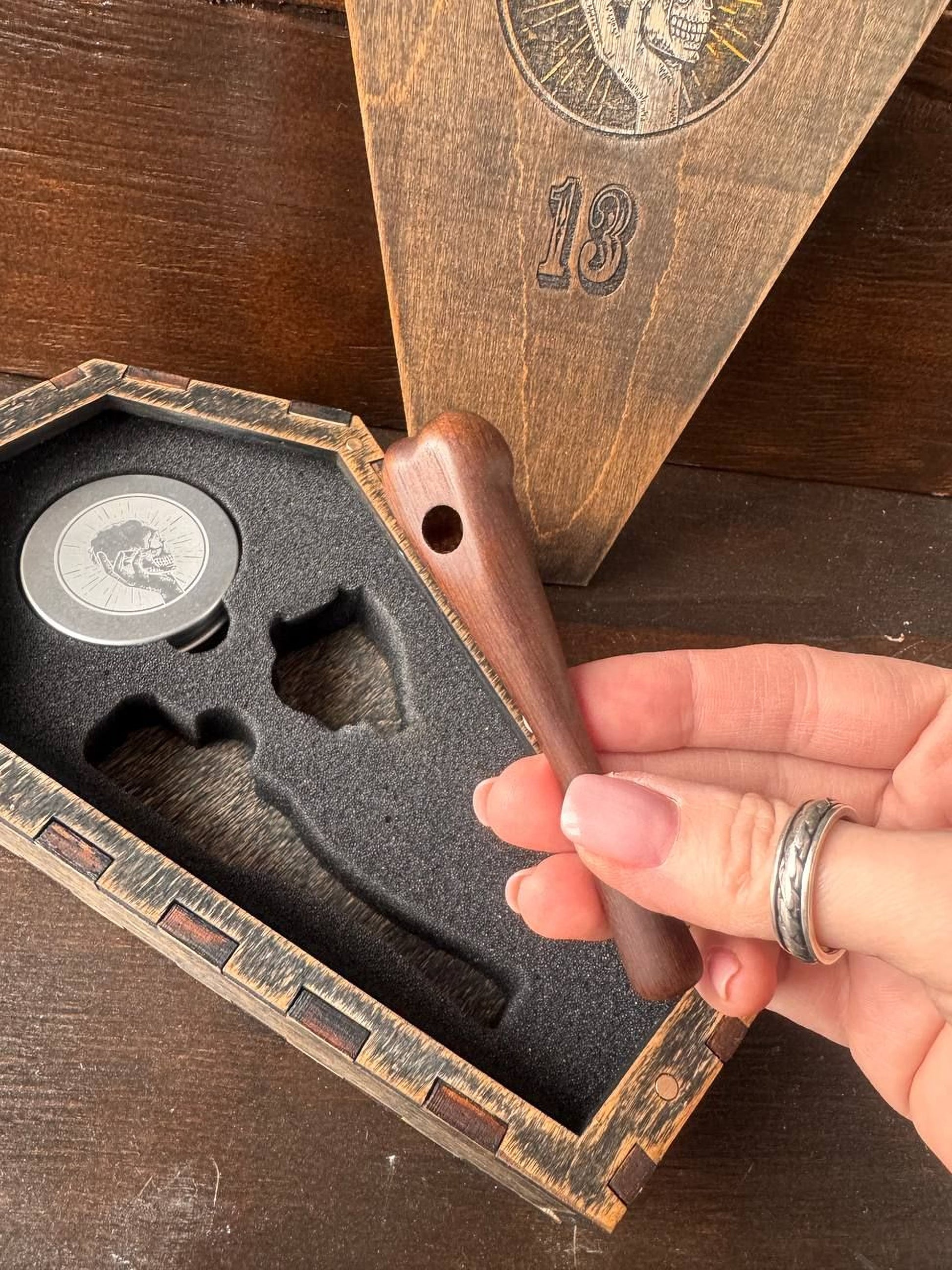 A hand holding a wooden object, possibly a tool or handle, next to a wooden box with a metal lid and a skull design.