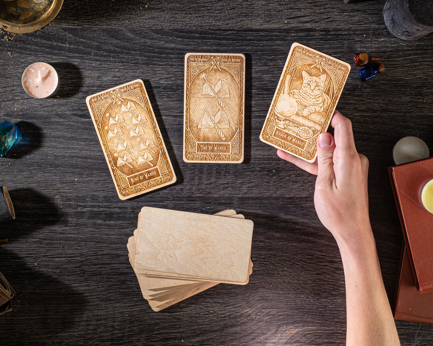 a person's hand holding three wooden tarot cards on a dark wooden surface, surrounded by various objects such as candles, books, and other items.