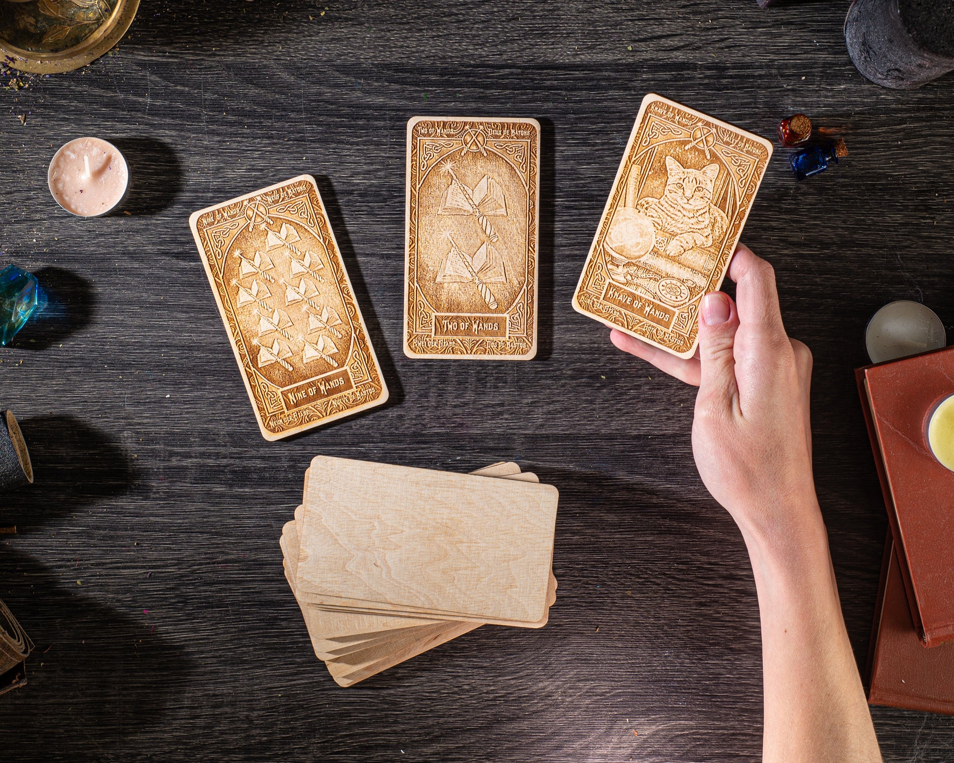 a person's hand holding three wooden tarot cards on a dark wooden surface, surrounded by various objects such as candles, books, and other items.