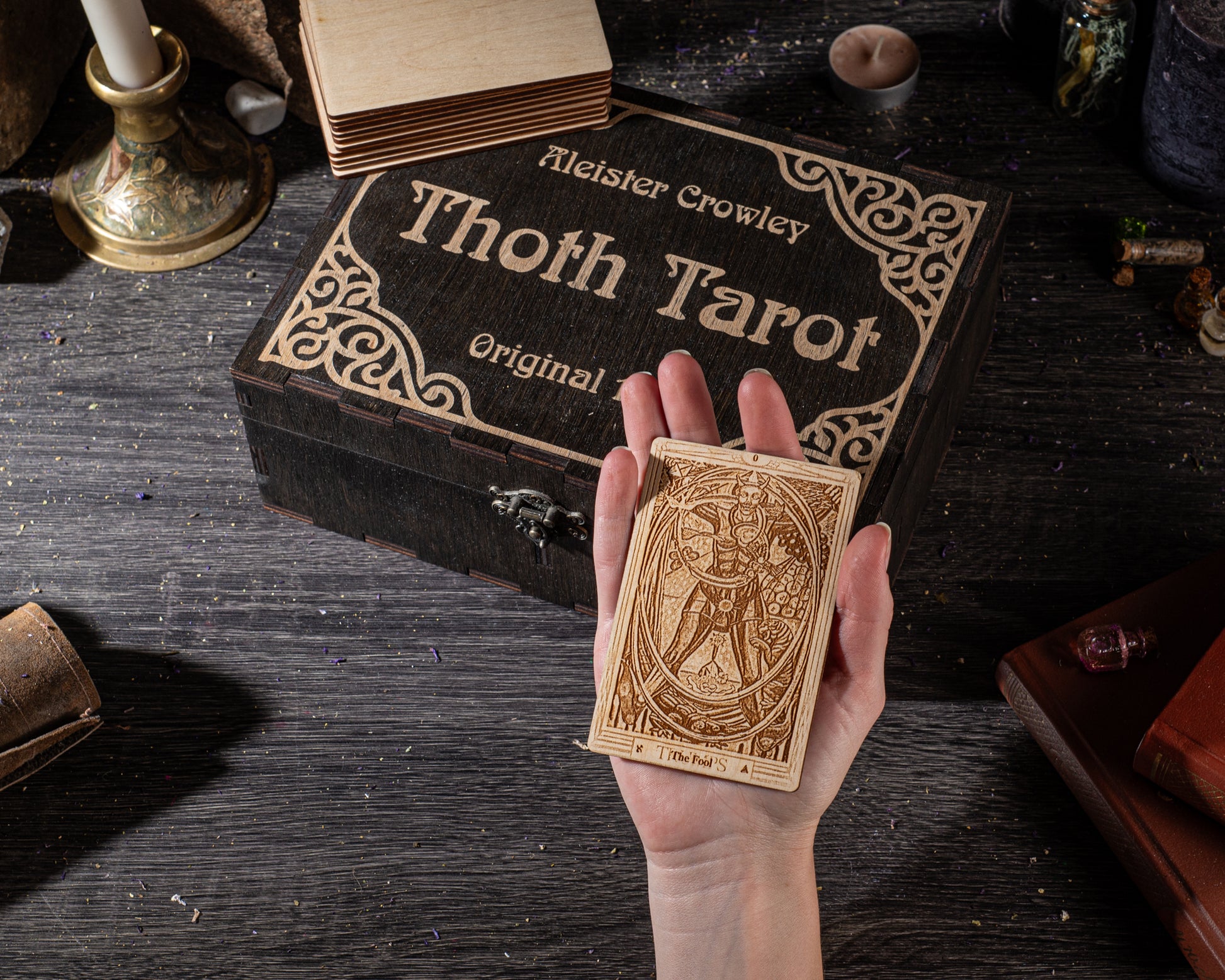 A hand holding a wooden tarot card in front of a wooden box with the name "Thoth Tarot" written on it.