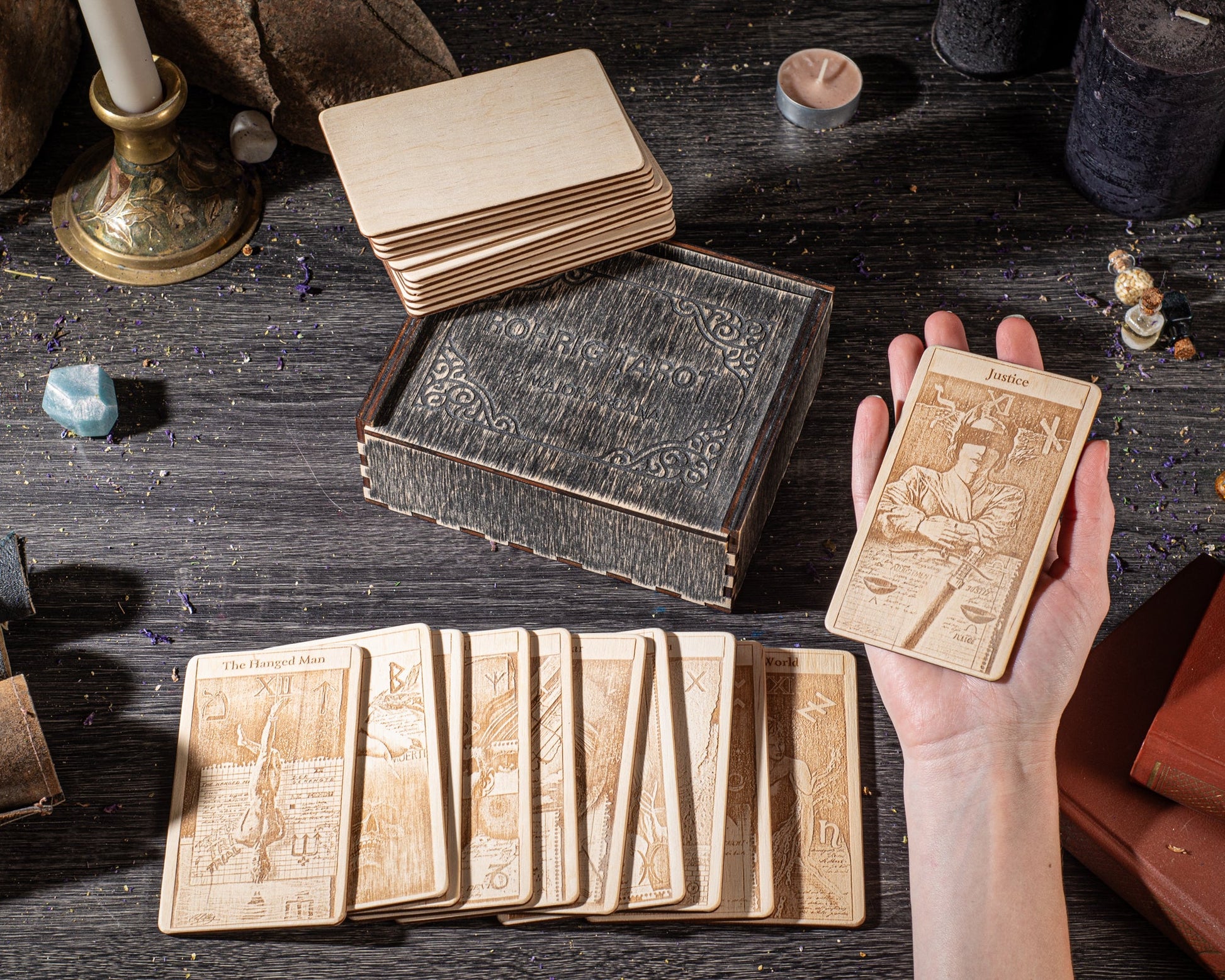 a hand holding a wooden tarot card, surrounded by a collection of wooden cards and a wooden box on a dark wooden surface.