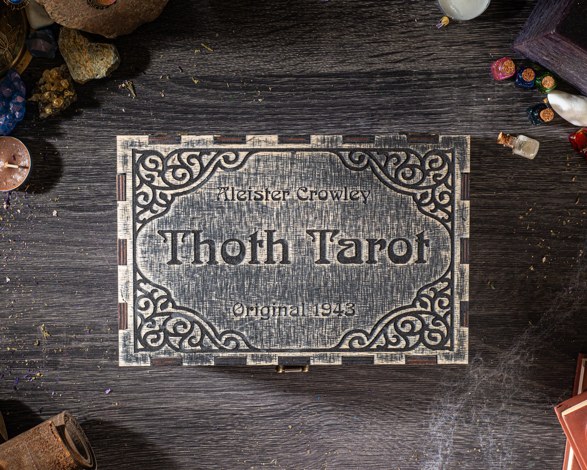A wooden box with a decorative border, featuring the name "Thoth Tarot" and the year "Original 1943" on it, placed on a wooden surface surrounded by various objects.