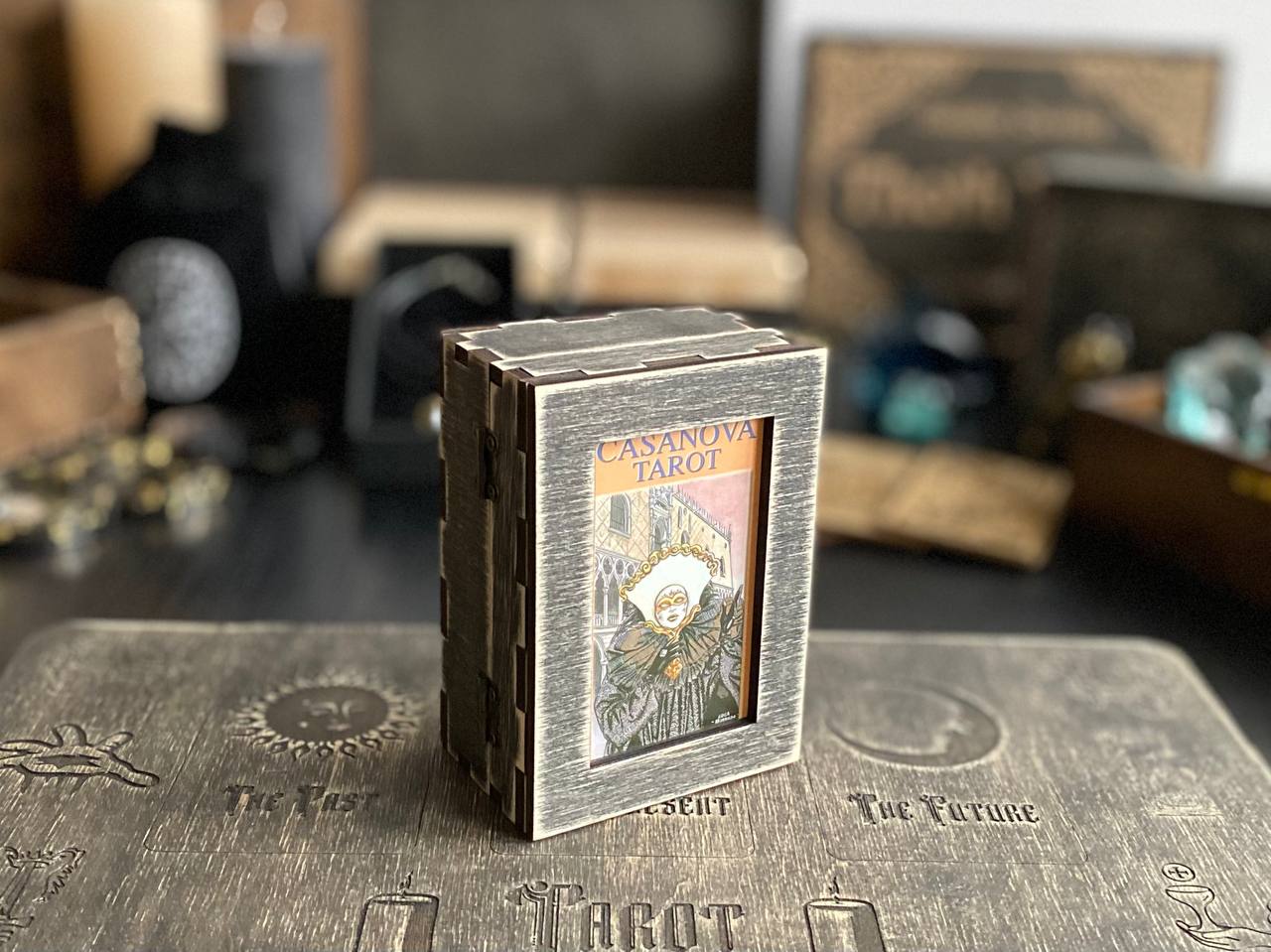 A small wooden box with a picture of a tarot card on it, placed on a wooden surface with various other objects in the background.