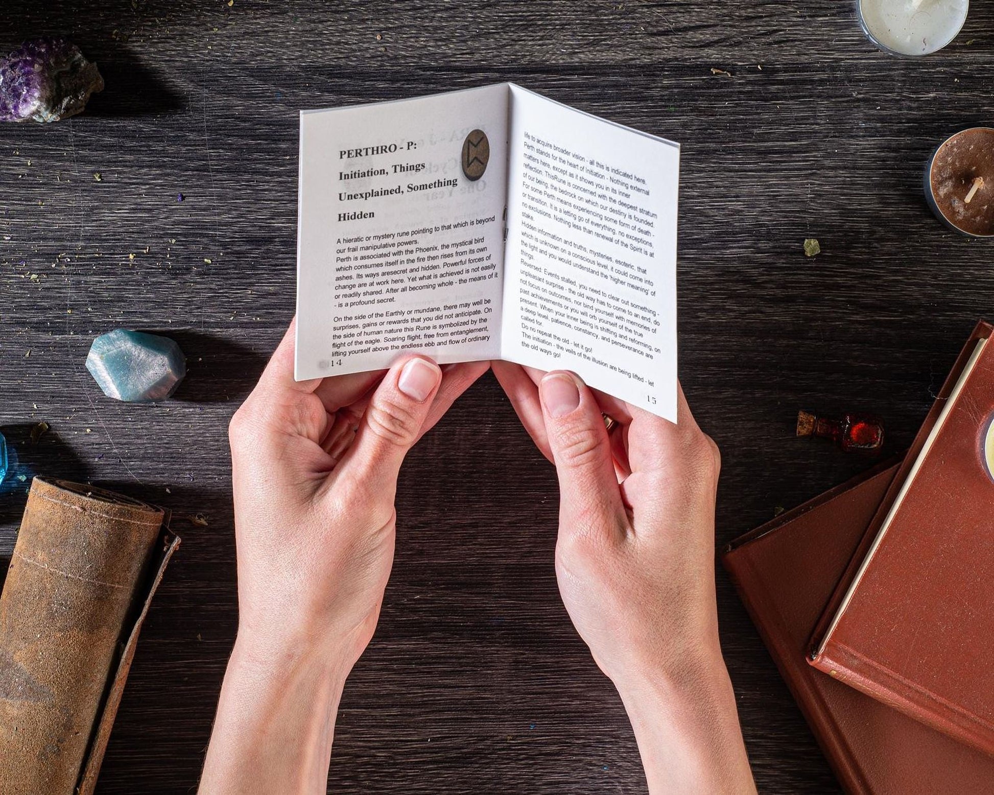 A person's hands are holding a book with text on the pages, which appear to be a book of spells or incantations, with various crystals and objects scattered around the wooden surface.