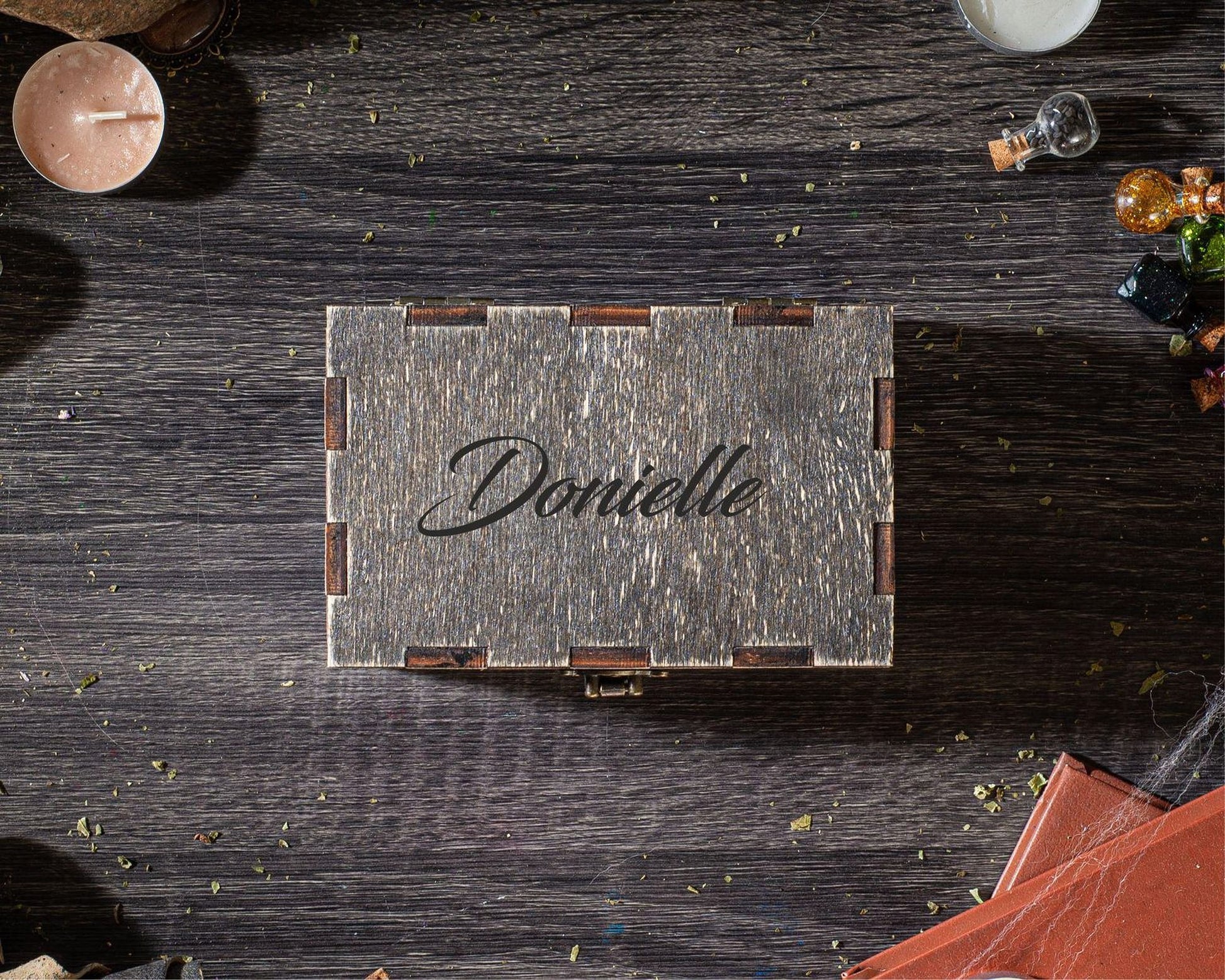 A wooden box with the name "Donielle" engraved on it, placed on a wooden surface surrounded by various objects such as a candle, a small glass bottle, and some small items.