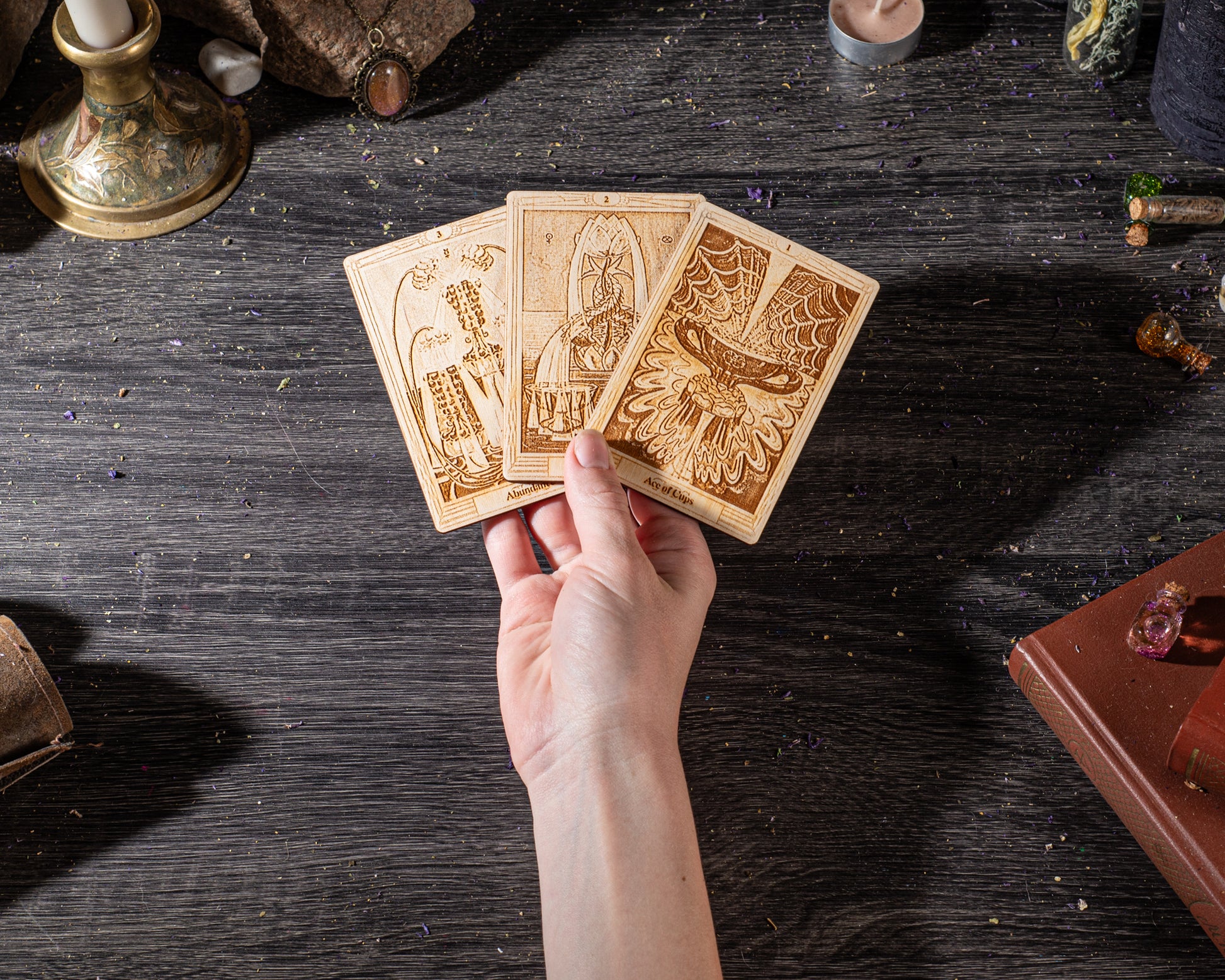 A hand is holding three tarot cards on a wooden surface, surrounded by various objects such as a candle, a book, and other items.