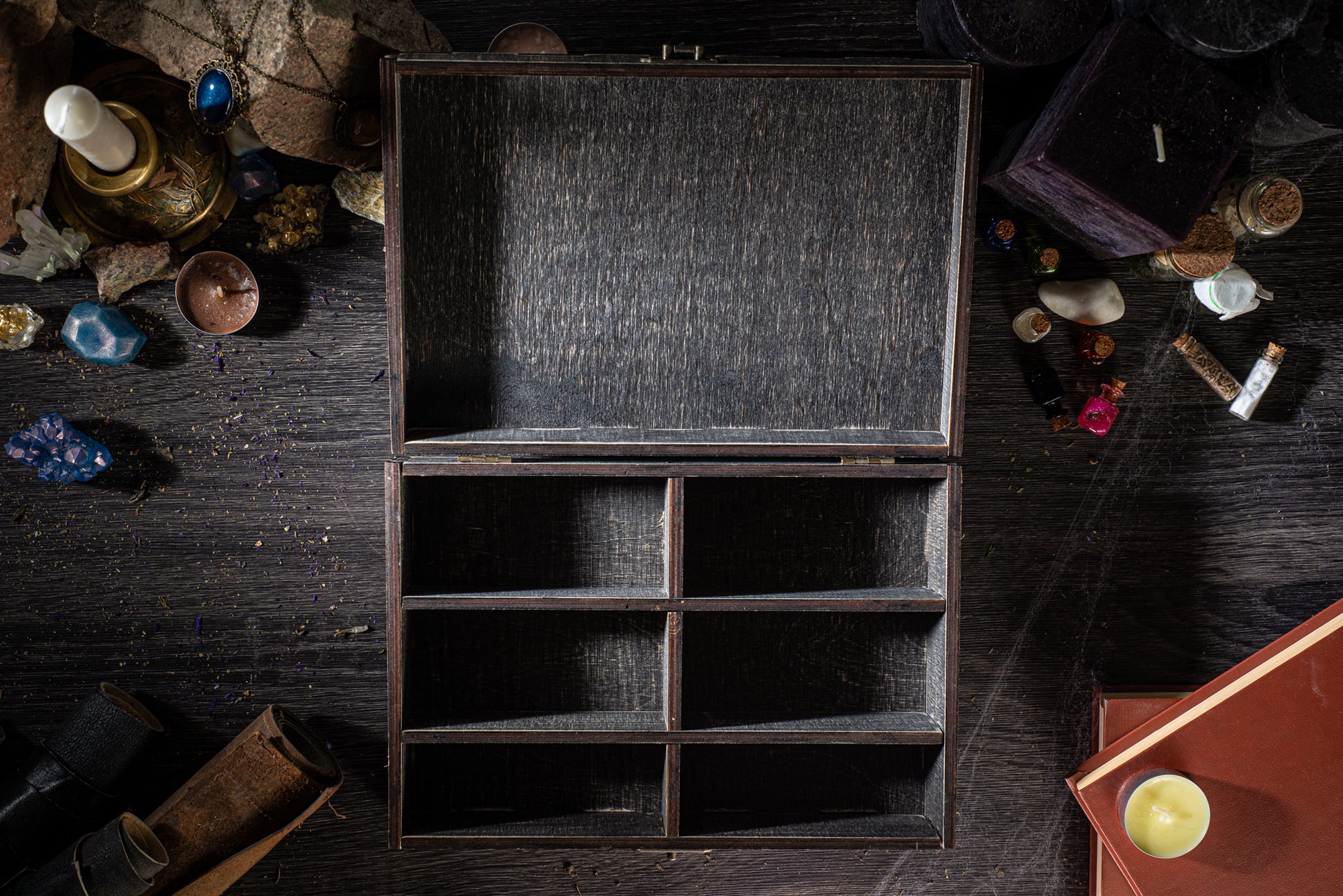 a wooden box with three compartments, surrounded by various objects such as candles, crystals, and books on a dark wooden surface.