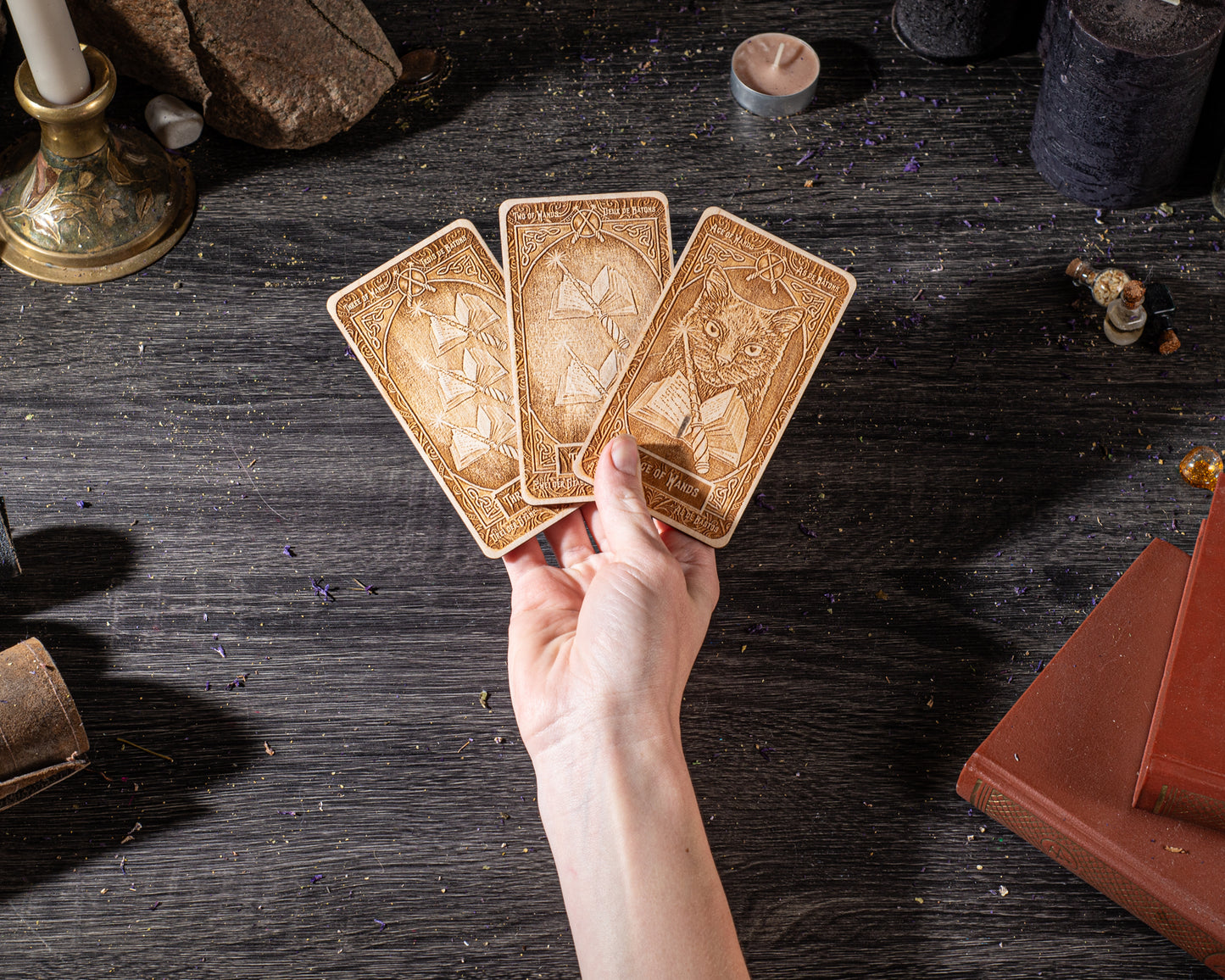 A hand is holding three tarot cards on a wooden surface, surrounded by various objects such as candles, books, and crystals.